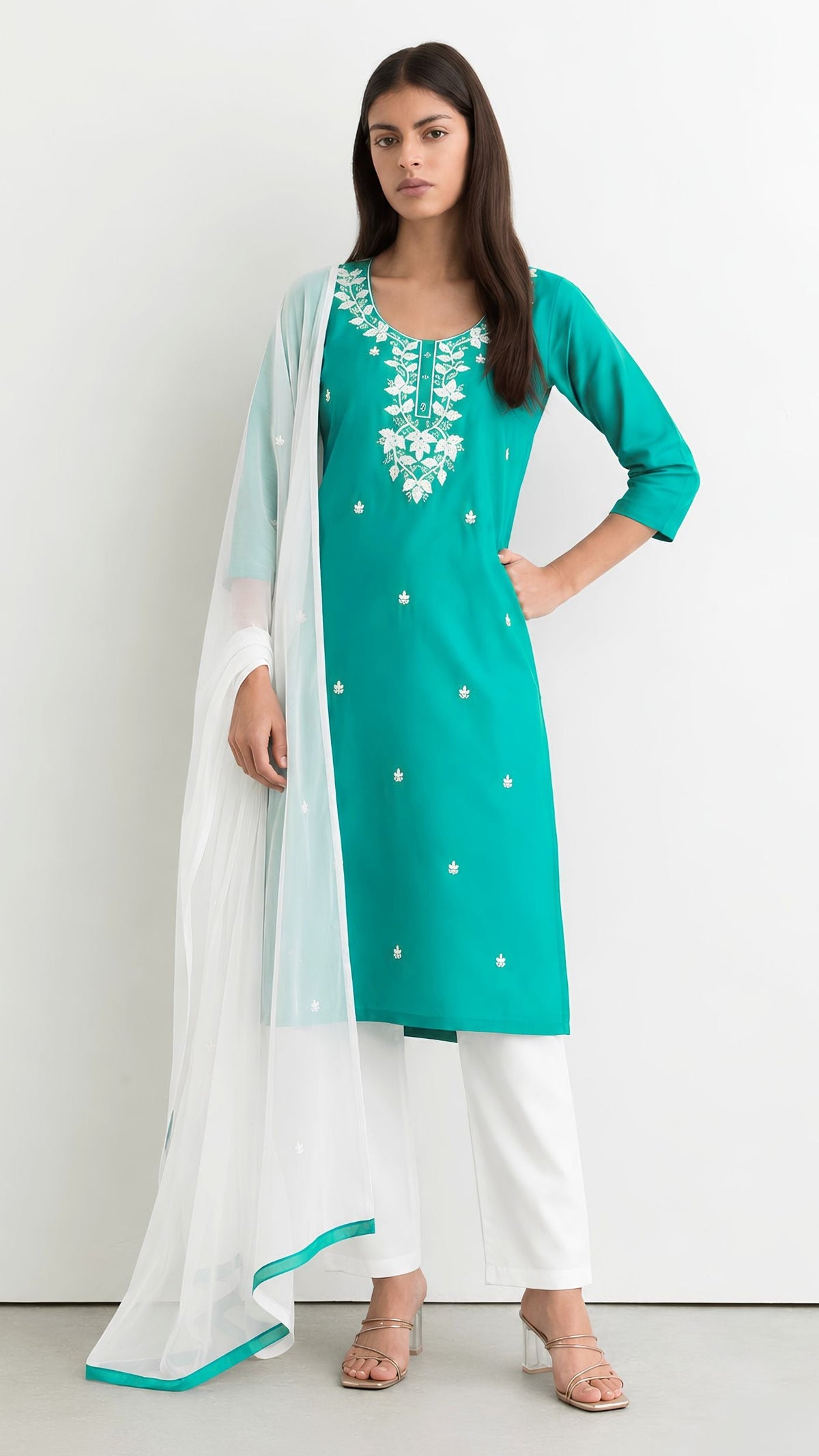 A woman modeling a teal green kurta with white floral embroidery on the neckline, paired with white pants and a sheer white dupatta with a teal border, set against a plain white studio background.
