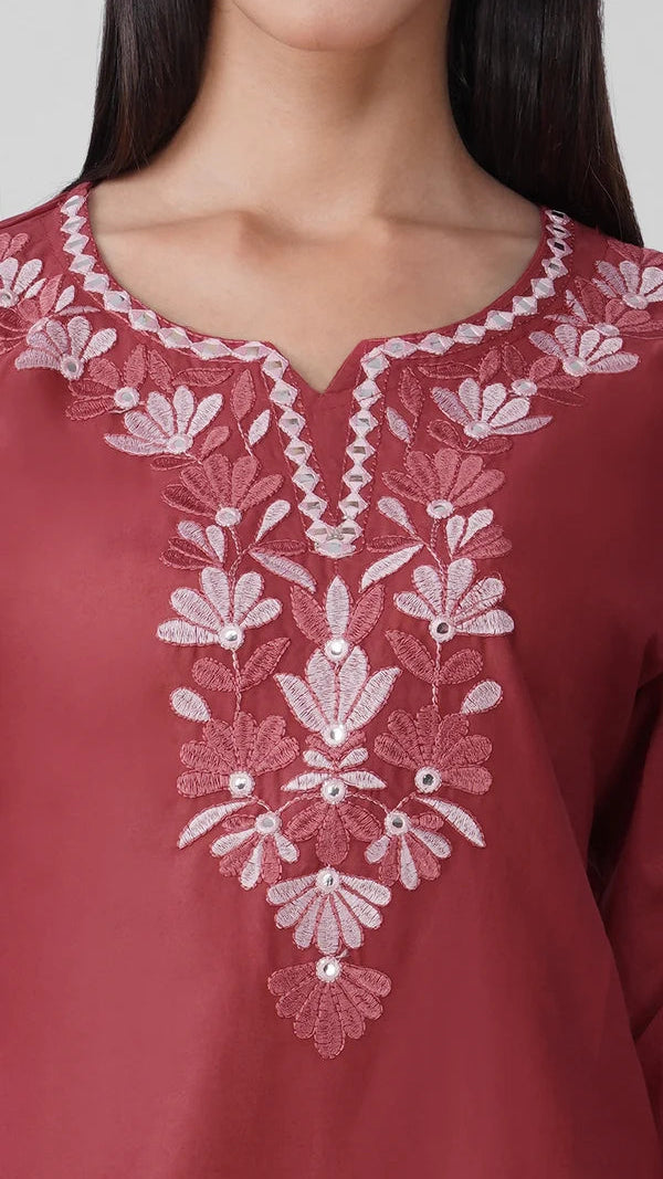 A close-up of a dusty rose pink kurta featuring intricate white floral embroidery and beadwork around the split neckline, worn by a model.