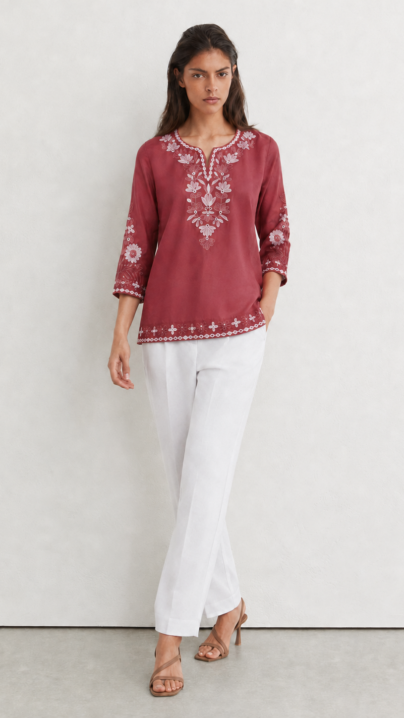 A woman modeling a dusty rose embroidered tunic top with white three-quarter sleeves and white pants, standing against a light gray textured studio wall.