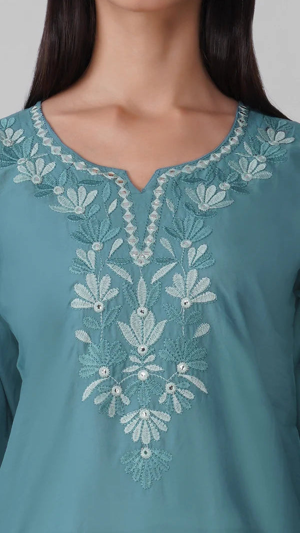 A close-up of a woman wearing a teal blue kurta with intricate white floral embroidery around the split neckline and down the center front.