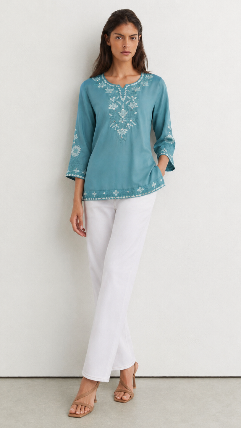 A woman modeling a teal blue embroidered tunic top with white three-quarter sleeves and white pants, standing against a plain white wall.