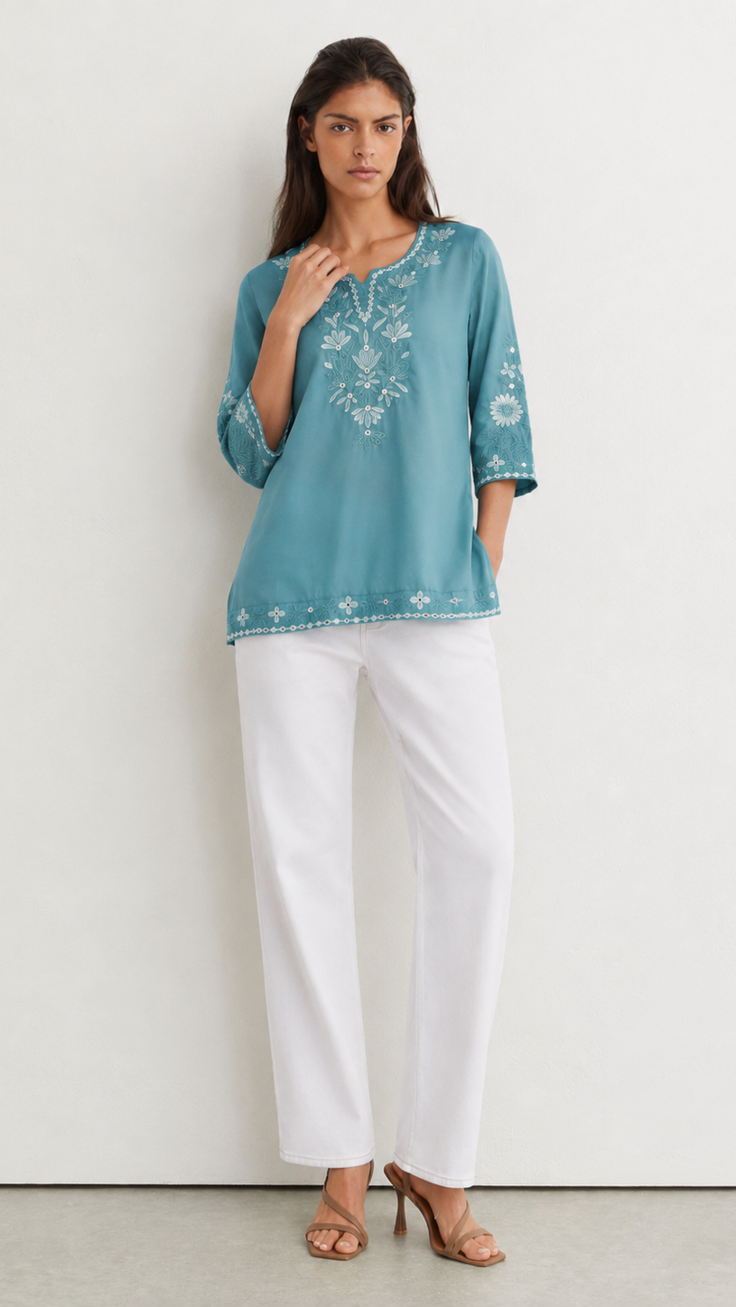 A woman modeling a turquoise blue embroidered tunic top with white three-quarter sleeves and white wide-leg trousers, standing against a plain white wall.