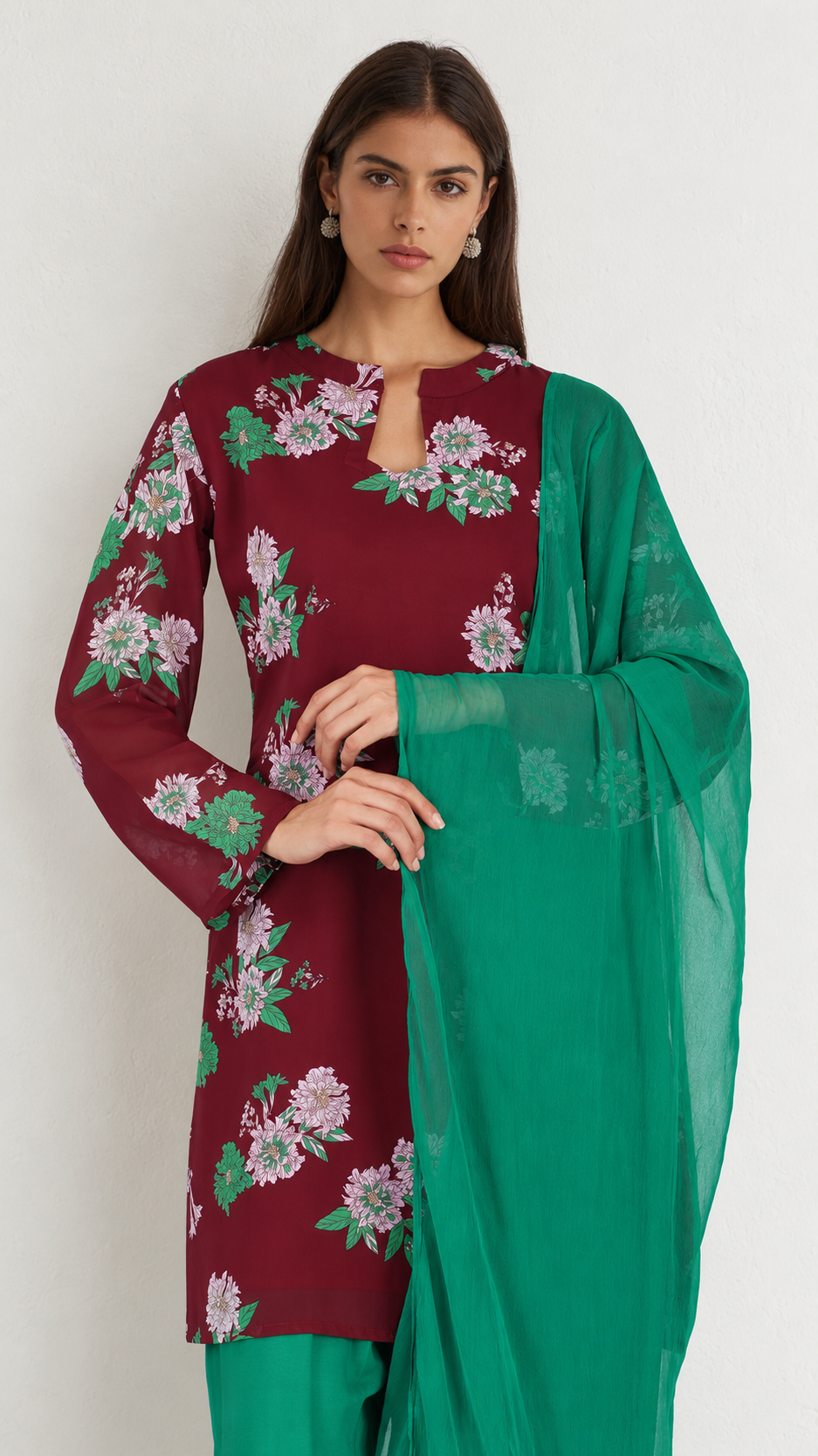 A woman modeling a traditional three-piece salwar kameez set. The kurta is deep maroon with a floral print in pink, green, and white, paired with a solid green dupatta and matching green trousers.