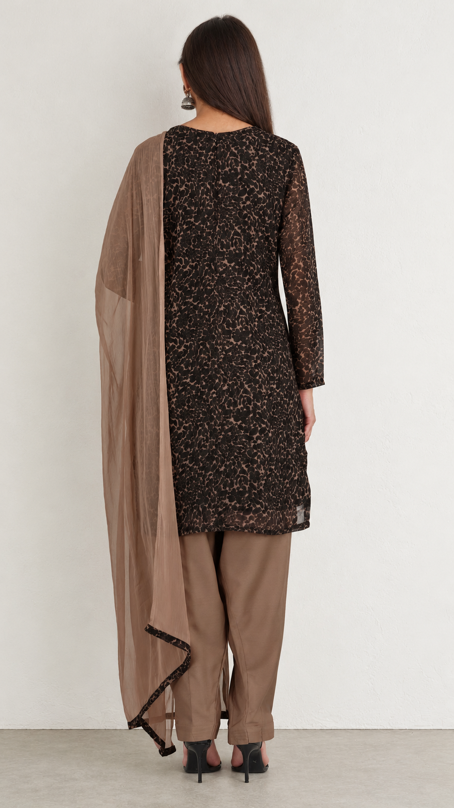 A woman shown from the back wearing a dark brown and black patterned kurta with sheer sleeves, paired with beige trousers and a matching beige dupatta, against a plain light gray wall.