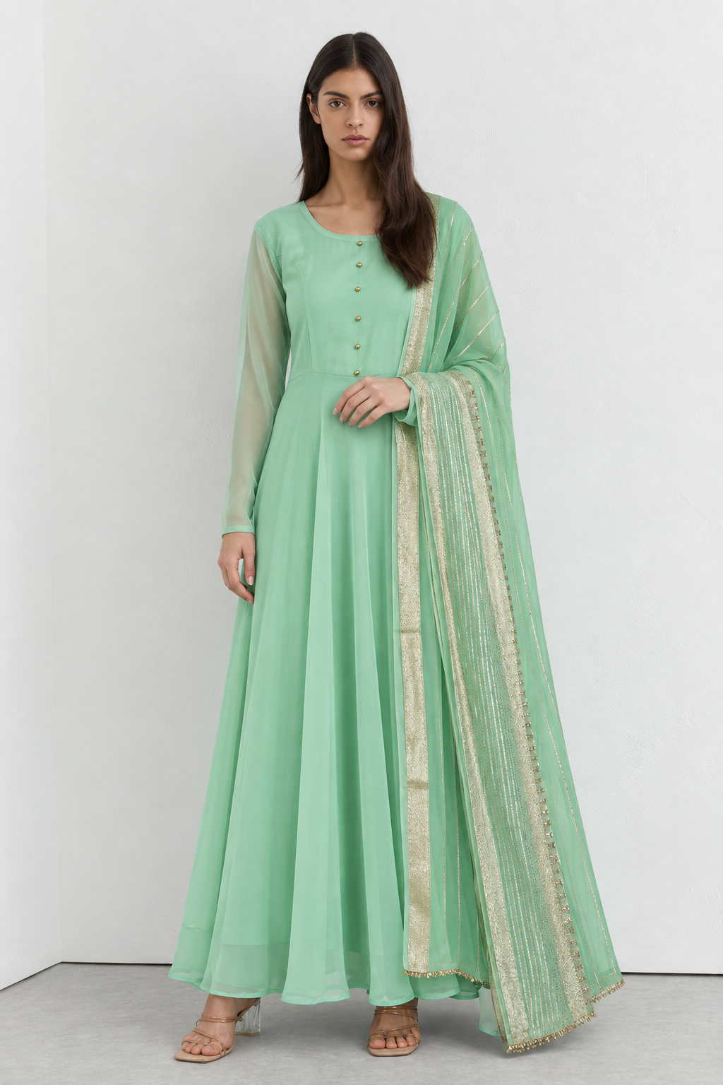 A woman modeling a mint green anarkali-style dress with a matching sheer dupatta featuring gold embroidery, set against a plain light gray studio background.