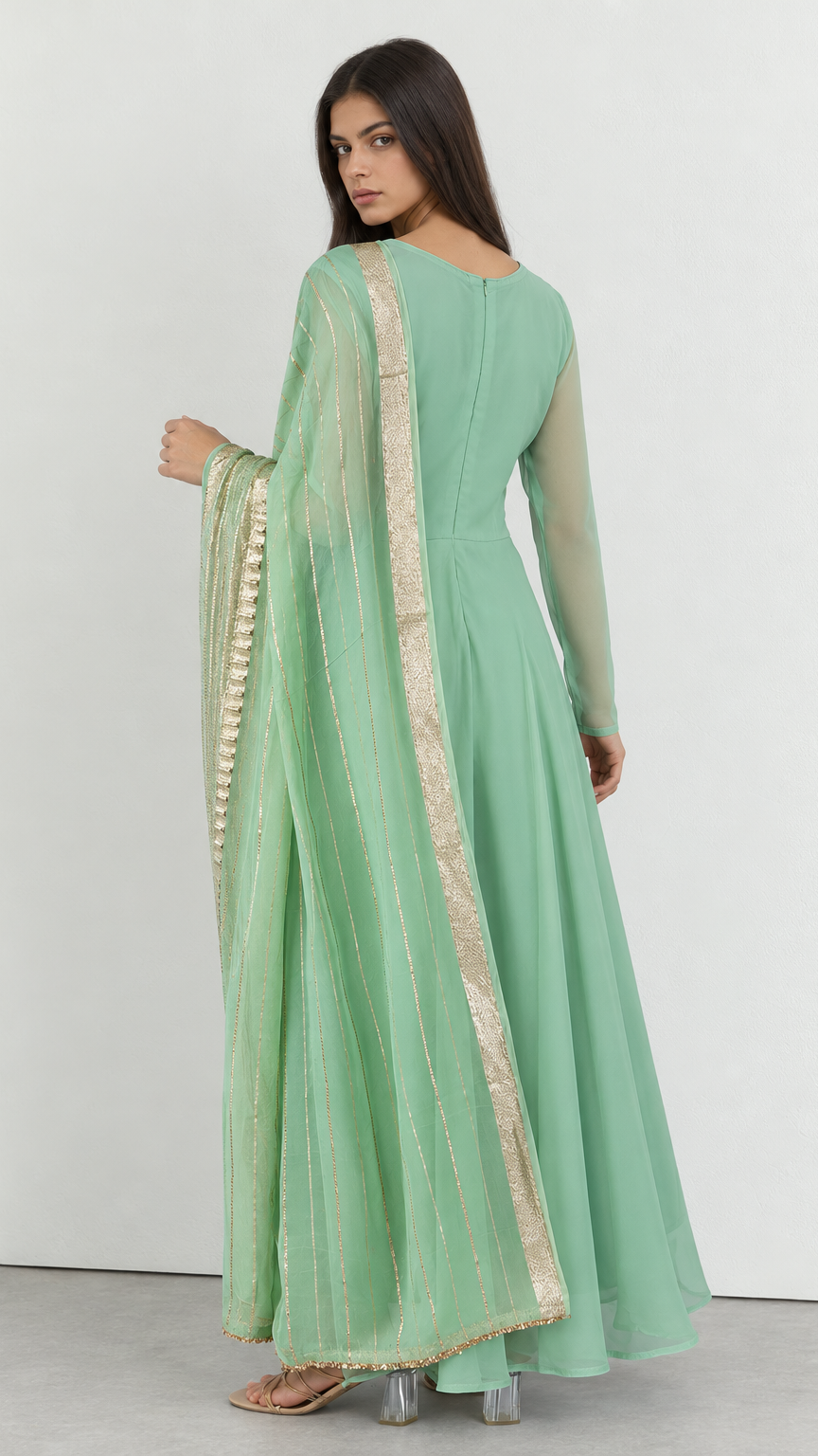 A woman modeling a mint green long-sleeve Anarkali gown with a matching sheer dupatta featuring gold zari stripes and border detailing, shown from the back against a plain white studio background.