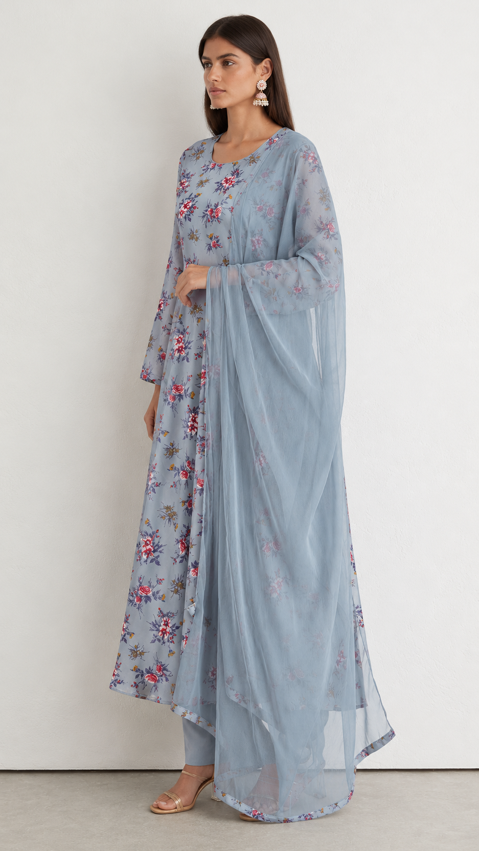 A woman modeling a light blue floral printed traditional dress with a matching sheer dupatta, standing against a plain off-white wall.