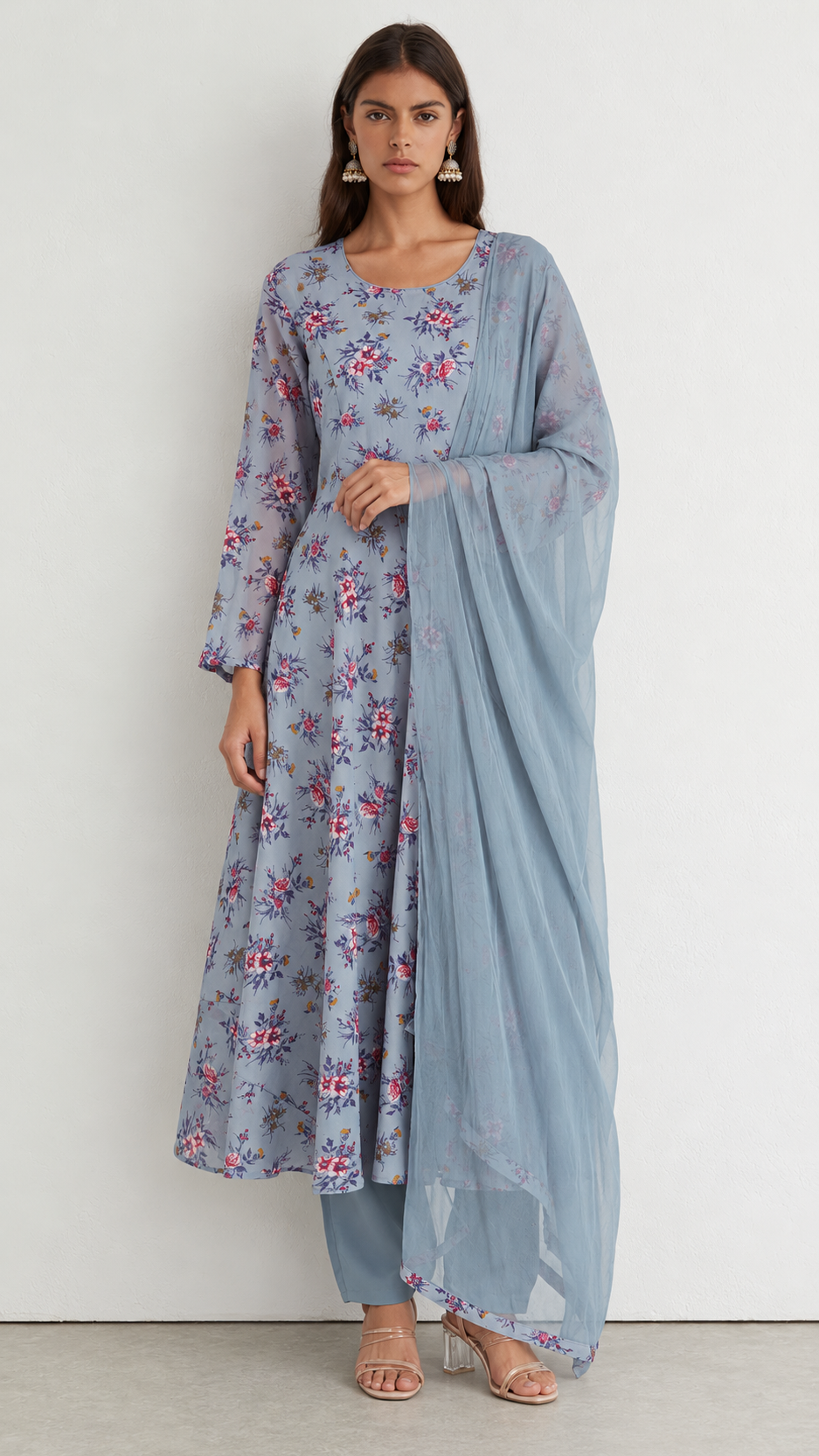 A woman modeling a light blue floral printed kurta set with matching pants and a sheer dupatta, standing against a plain off-white wall.