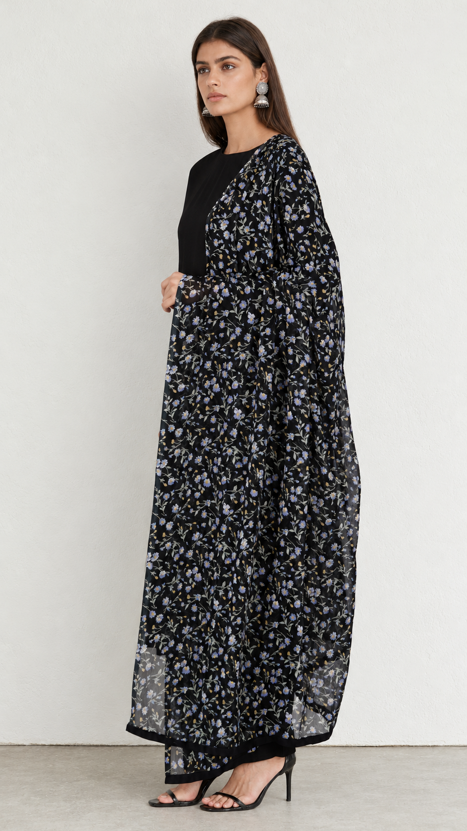A woman modeling a long black maxi dress with a dense white and gray floral print, styled with a solid black long-sleeve top and black high-heeled sandals against a plain light gray wall.