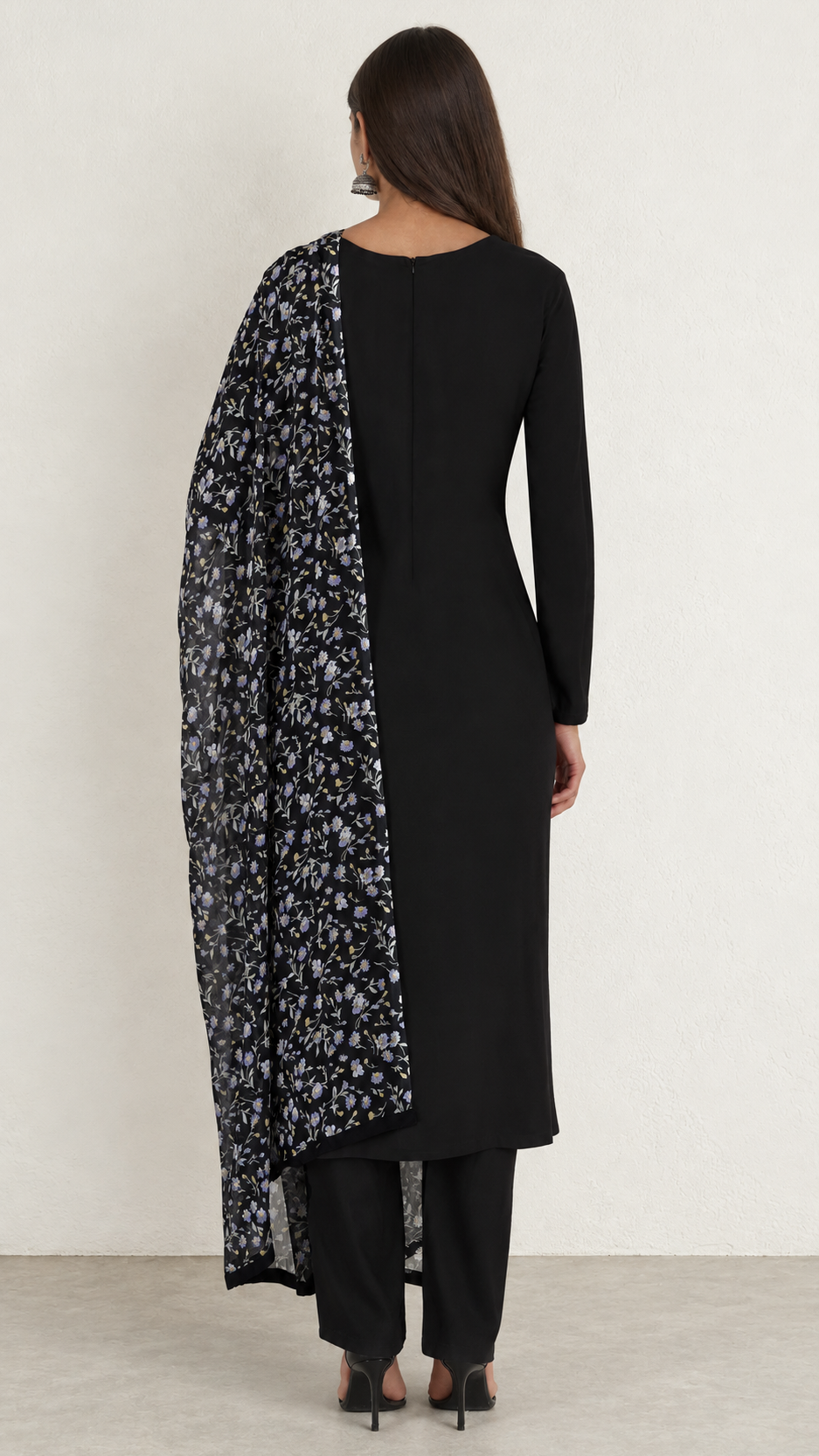 A woman shown from the back wearing a black long kurta with a black and white floral printed dupatta draped over one shoulder, paired with black pants and black heels, against a plain light gray studio background.