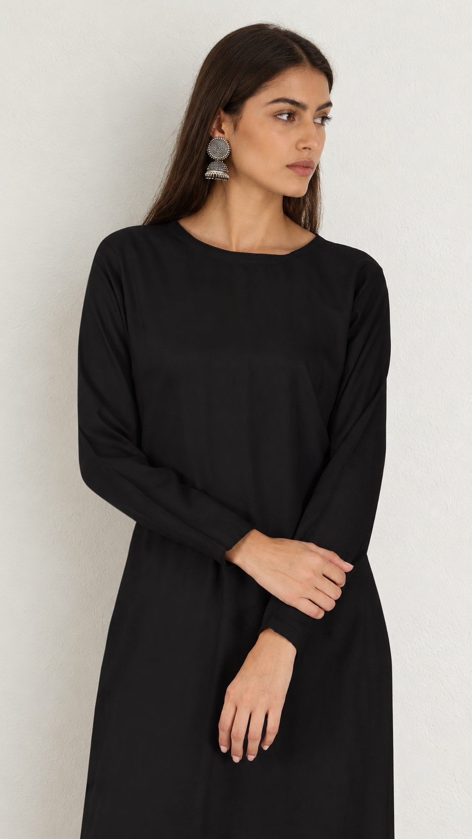 A woman wearing a long-sleeved black dress with a boat neckline, standing against a plain light gray wall.