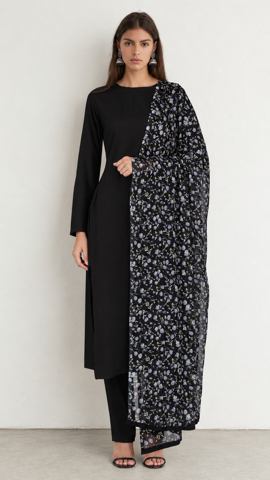 A woman modeling a black long kurta with a black and white floral printed dupatta, standing against a plain off-white wall.