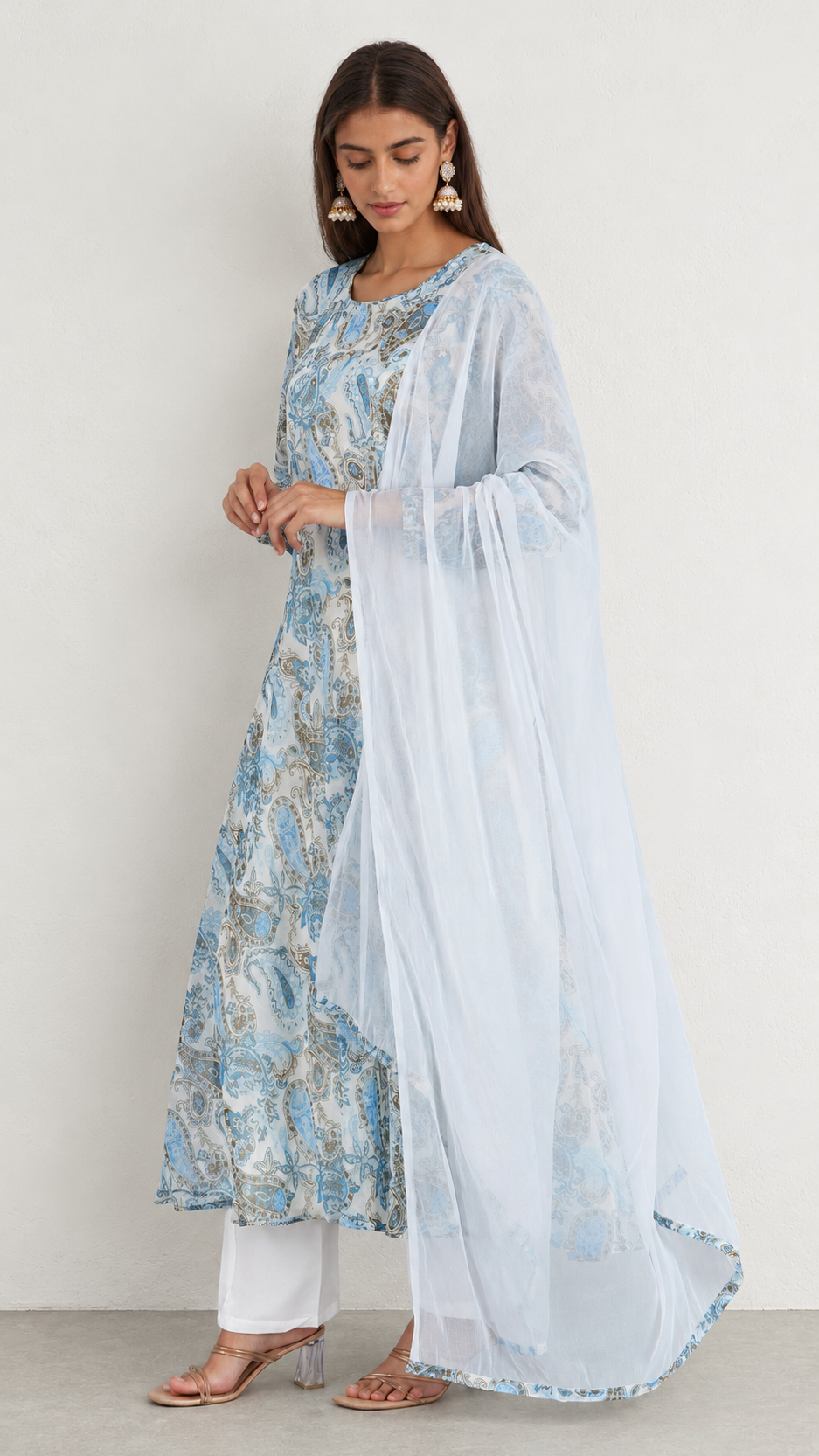 A woman models a traditional Indian outfit consisting of a light blue and white printed kurta, white pants, and a sheer light blue dupatta, set against a plain off-white studio background.