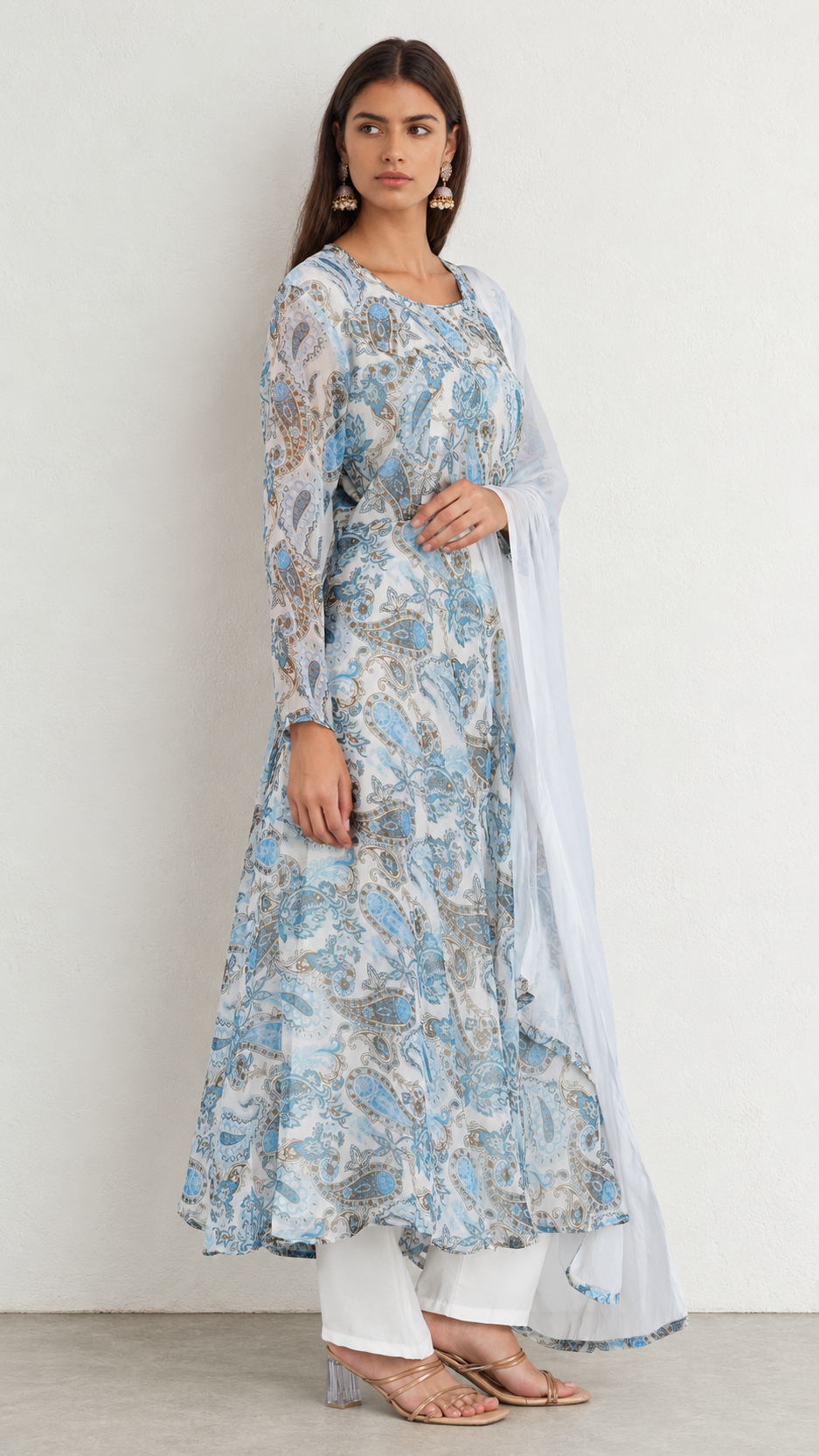 A woman modeling a light blue and white printed kurta set with a white dupatta, white pants, and clear high-heeled sandals against a plain off-white wall.