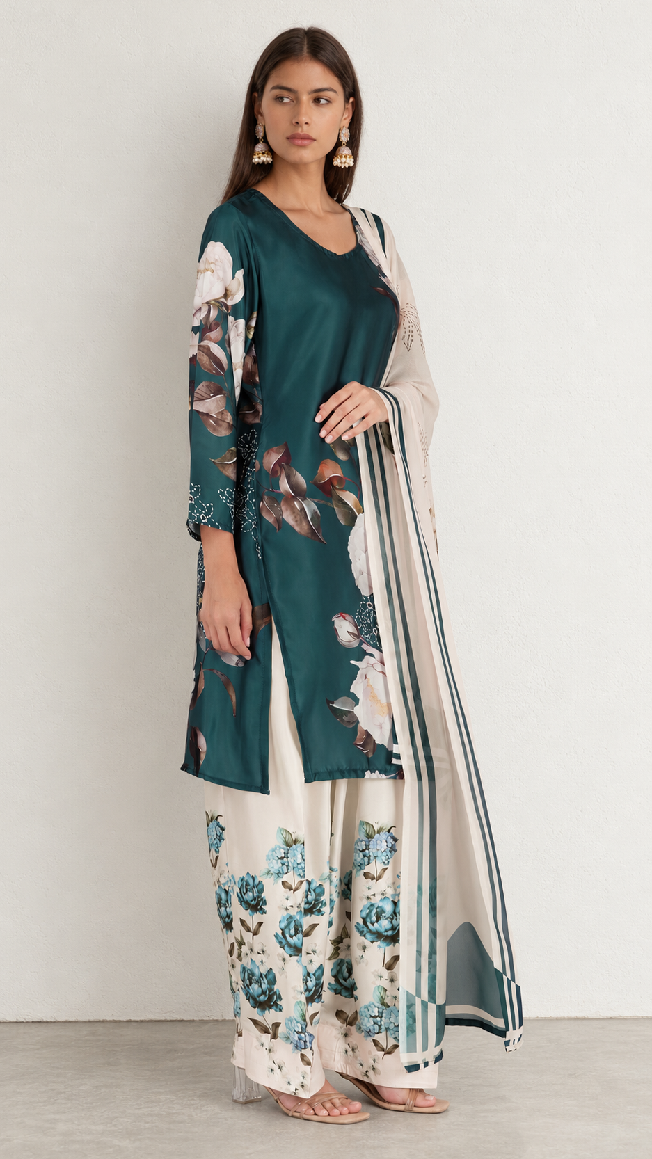 A woman modeling a traditional Indian outfit consisting of a dark teal green kurta with floral prints, wide-leg cream pants with blue floral patterns, and a sheer cream dupatta with green stripes.