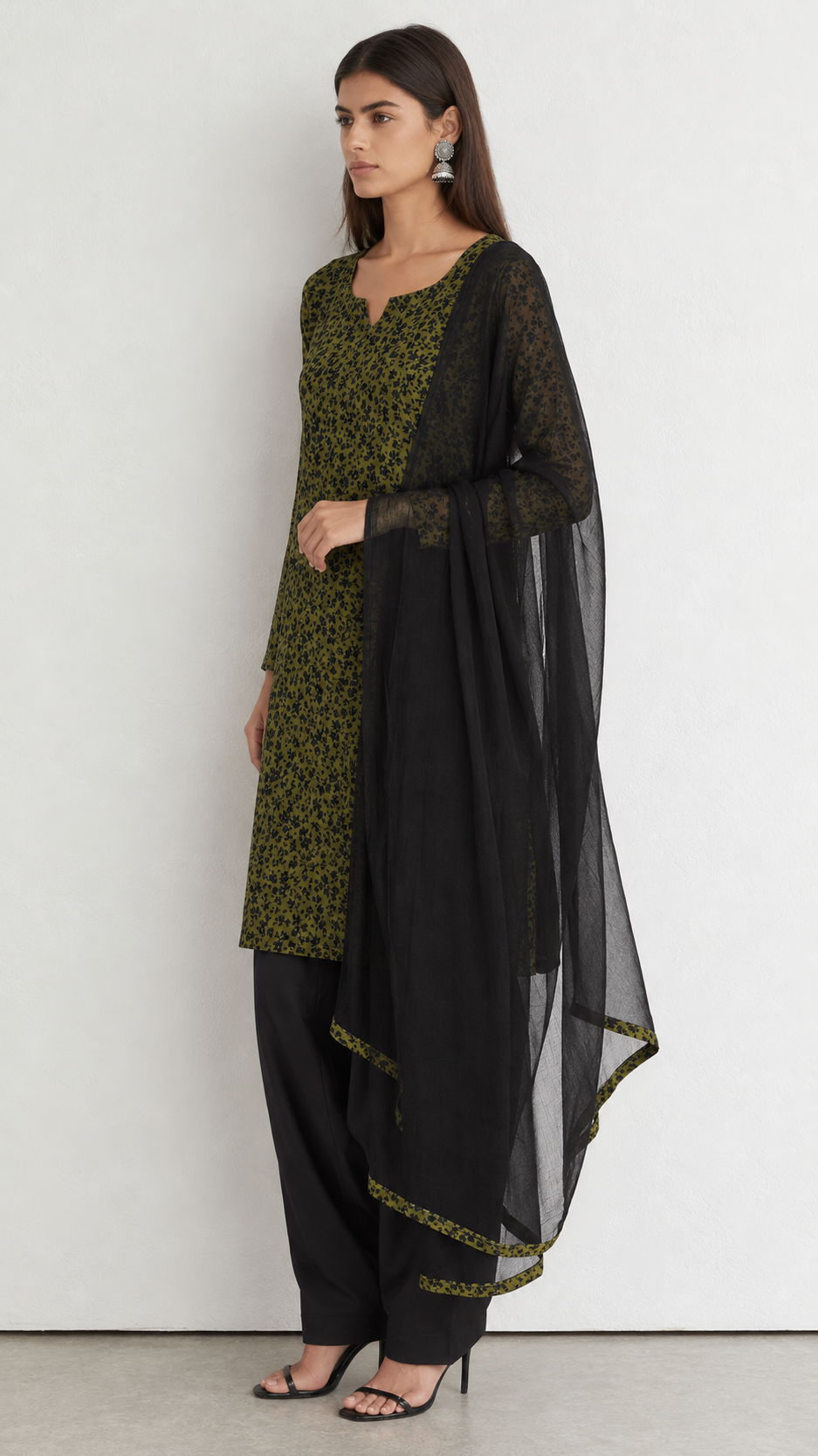 A woman modeling a black and olive green ethnic suit set, featuring a patterned kurta, black pants, and a sheer black dupatta, set against a plain light gray studio background.