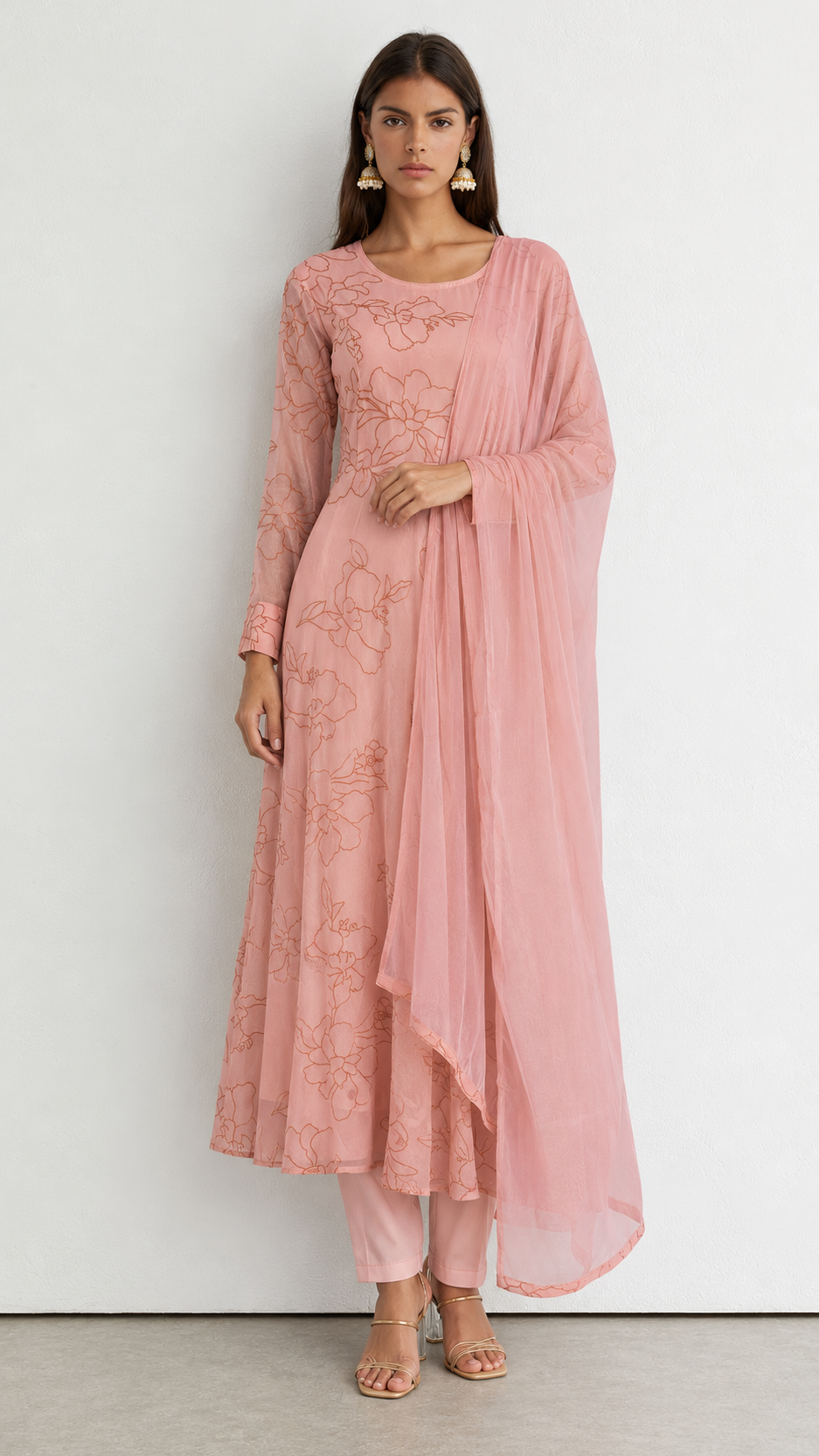 A woman modeling a dusty pink traditional Indian outfit consisting of a long-sleeved embroidered kurta, matching trousers, and a sheer draped scarf, set against a plain white wall.