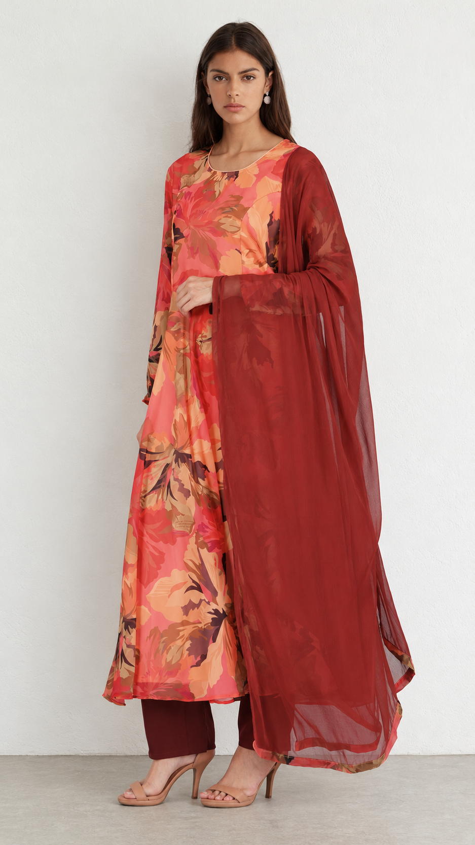 A woman models a traditional Indian outfit consisting of a floral printed kurta in shades of orange, pink, and red, paired with matching red pants and a sheer red dupatta, set against a plain white studio background.