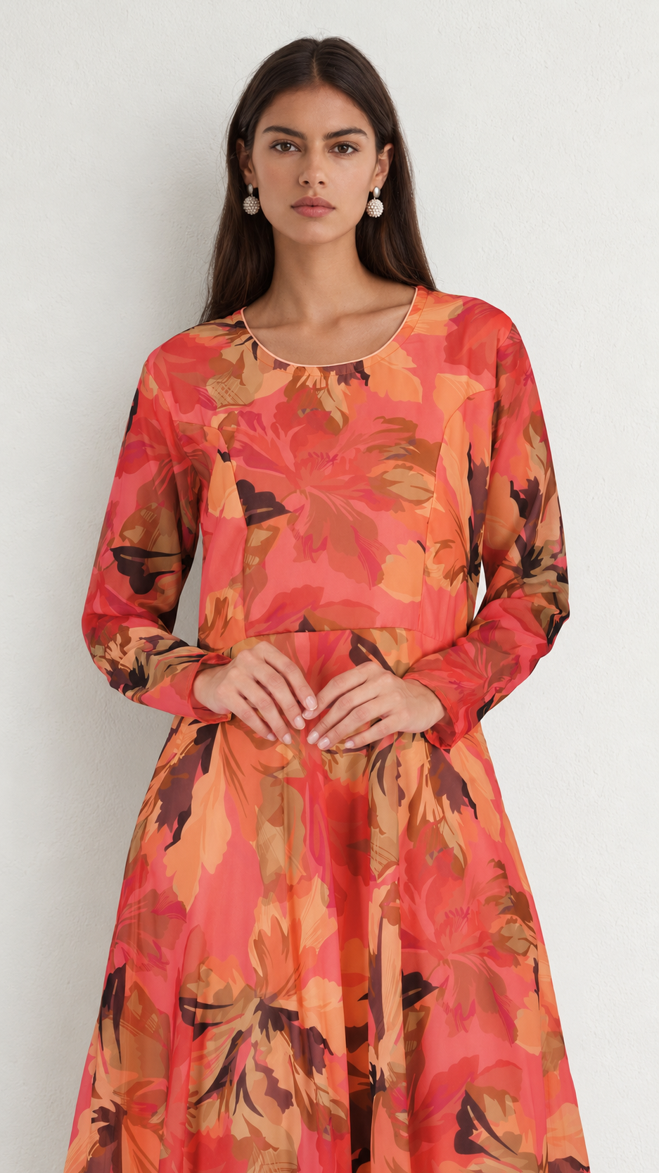 A woman modeling a long-sleeve, round-neck dress with a vibrant orange, red, and brown floral pattern against a plain light gray background.