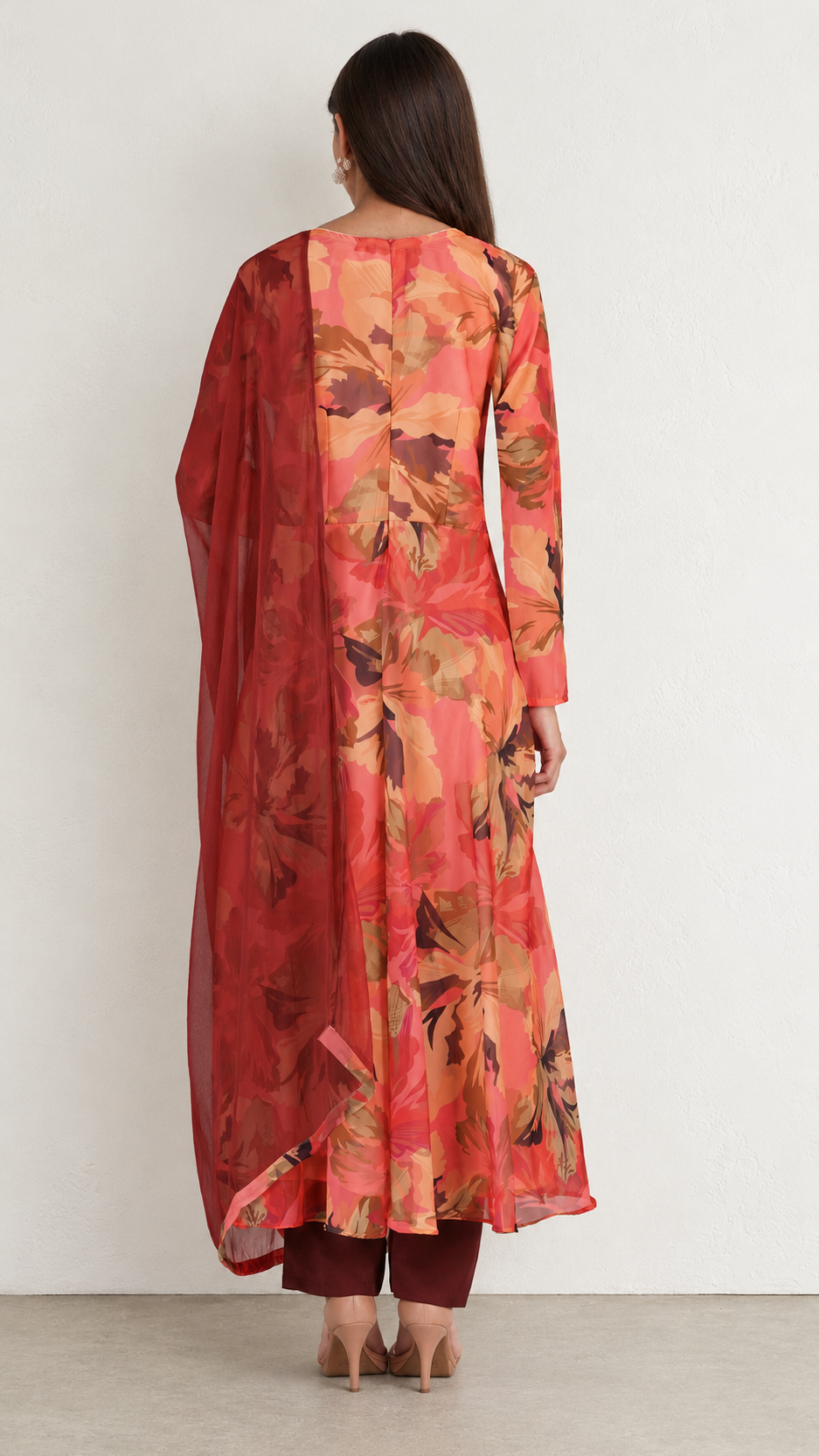 A woman viewed from the back wearing a long-sleeved floral kurta in red, orange, and brown tones, paired with a matching sheer dupatta and dark trousers, standing against a plain off-white wall.