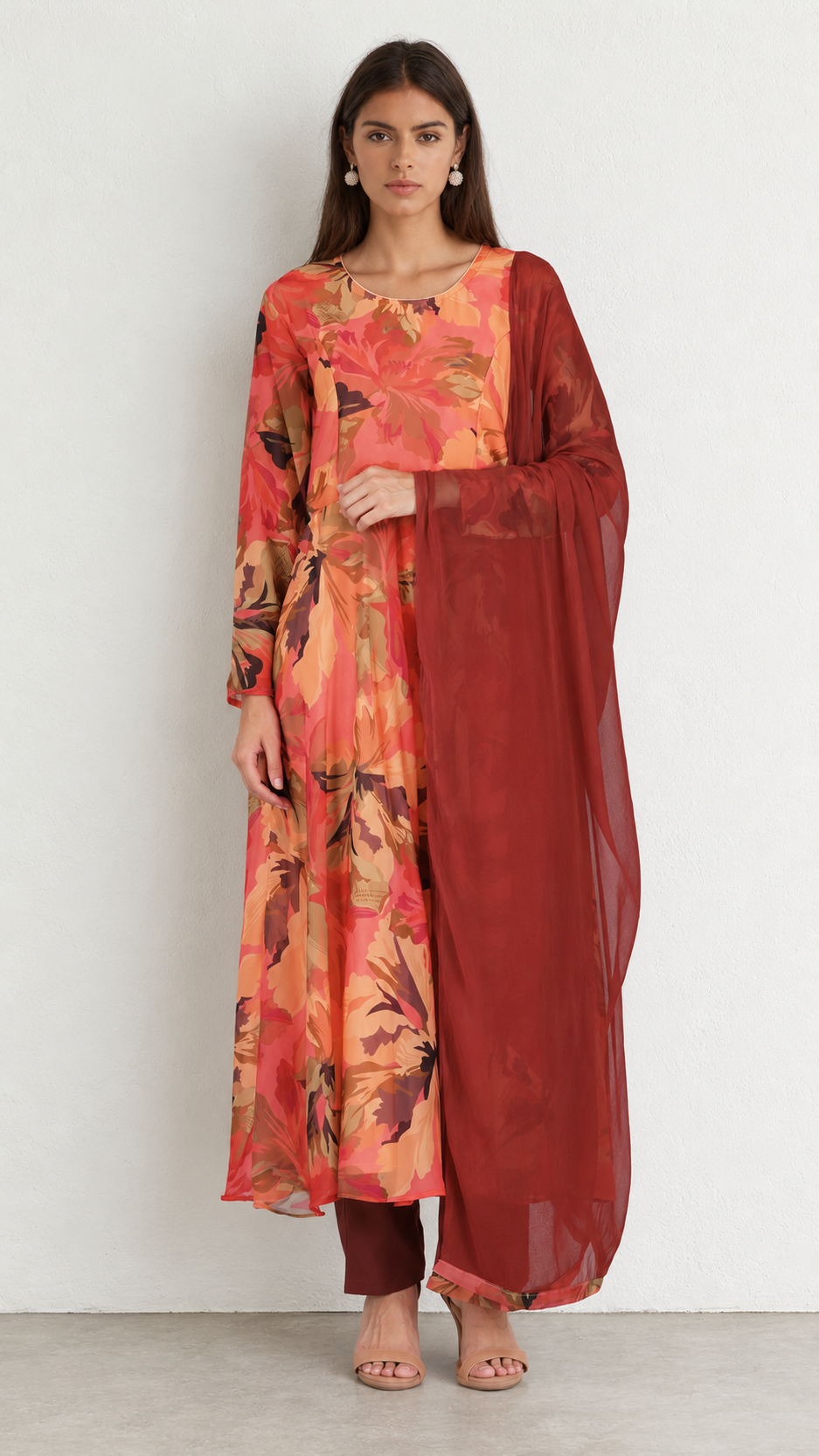 A woman modeling a traditional Indian salwar kameez set featuring a rust-orange and peach floral print kurta with a solid dark red dupatta, set against a plain white studio background.