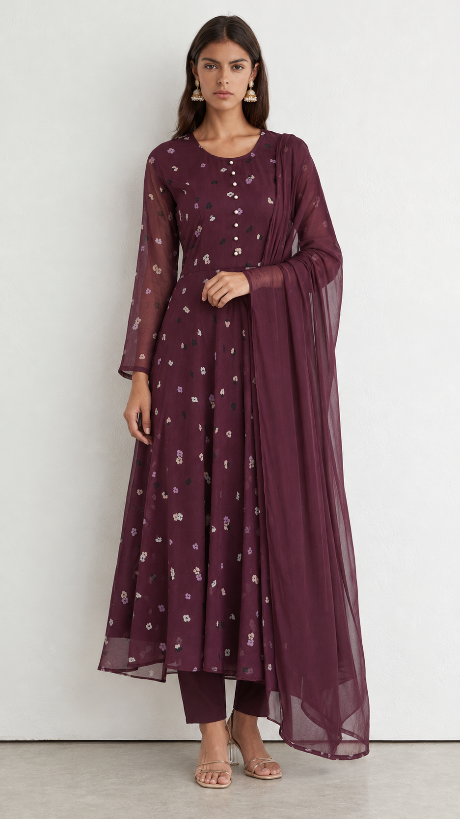 A woman modeling a deep plum purple traditional Indian outfit consisting of a long-sleeved kurta with small white floral prints, matching trousers, and a sheer matching dupatta, set against a plain light gray wall.