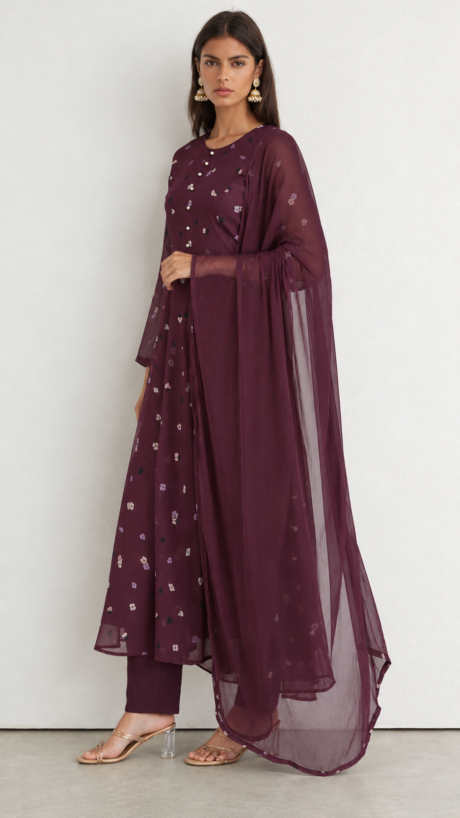 A woman models a deep plum purple traditional Indian outfit consisting of a kurta with floral embroidery, matching trousers, and a sheer dupatta, set against a plain light gray background.