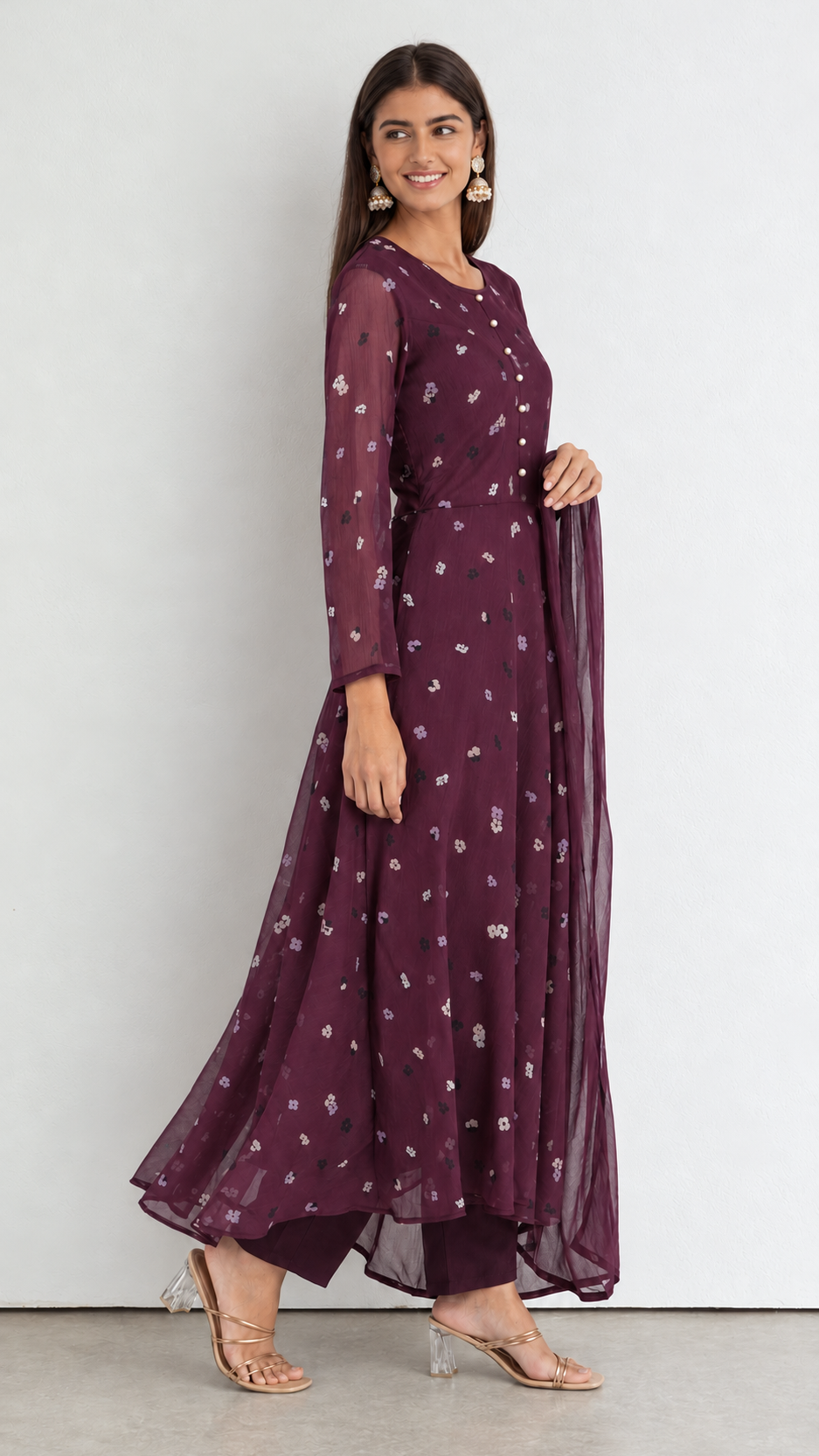 A woman modeling a long-sleeved, floor-length maroon anarkali kurta with white floral prints and a matching sheer dupatta, set against a plain light gray studio background.