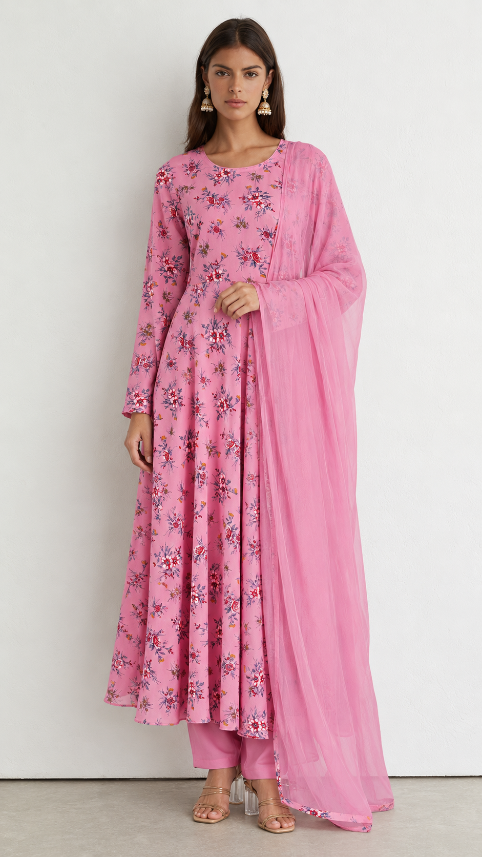 A woman modeling a pink long kurta set with floral embroidery and a matching sheer dupatta, standing against a plain light gray wall.
