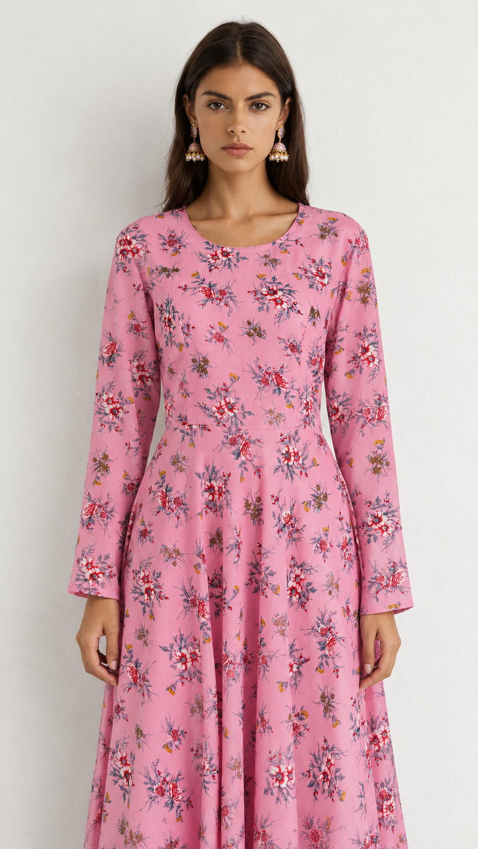 A woman wearing a long-sleeve pink maxi dress with a dense red, green, and white floral print, standing against a plain light gray background.