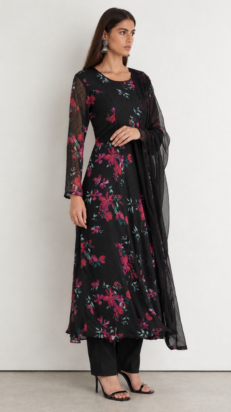 A woman modeling a black long kurta with a vibrant pink and red floral print, paired with black pants and a sheer black dupatta, set against a plain light gray studio background.