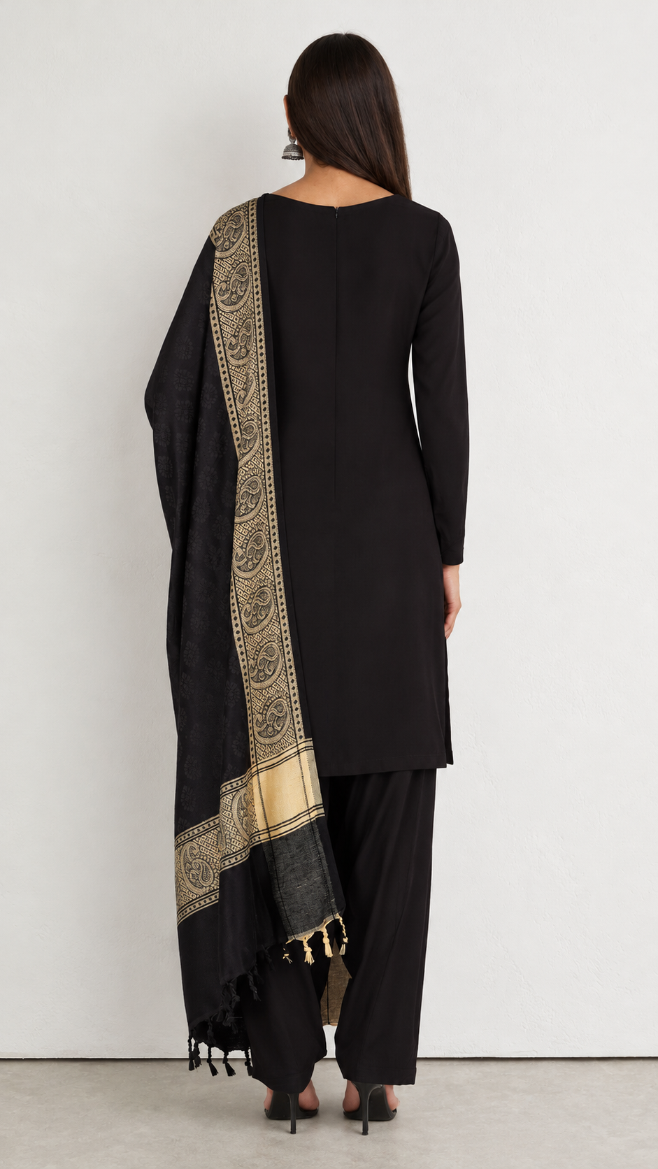A woman shown from the back wearing a black long kurta and wide-leg pants with a black dupatta featuring a prominent gold and beige patterned border, set against a plain white wall.