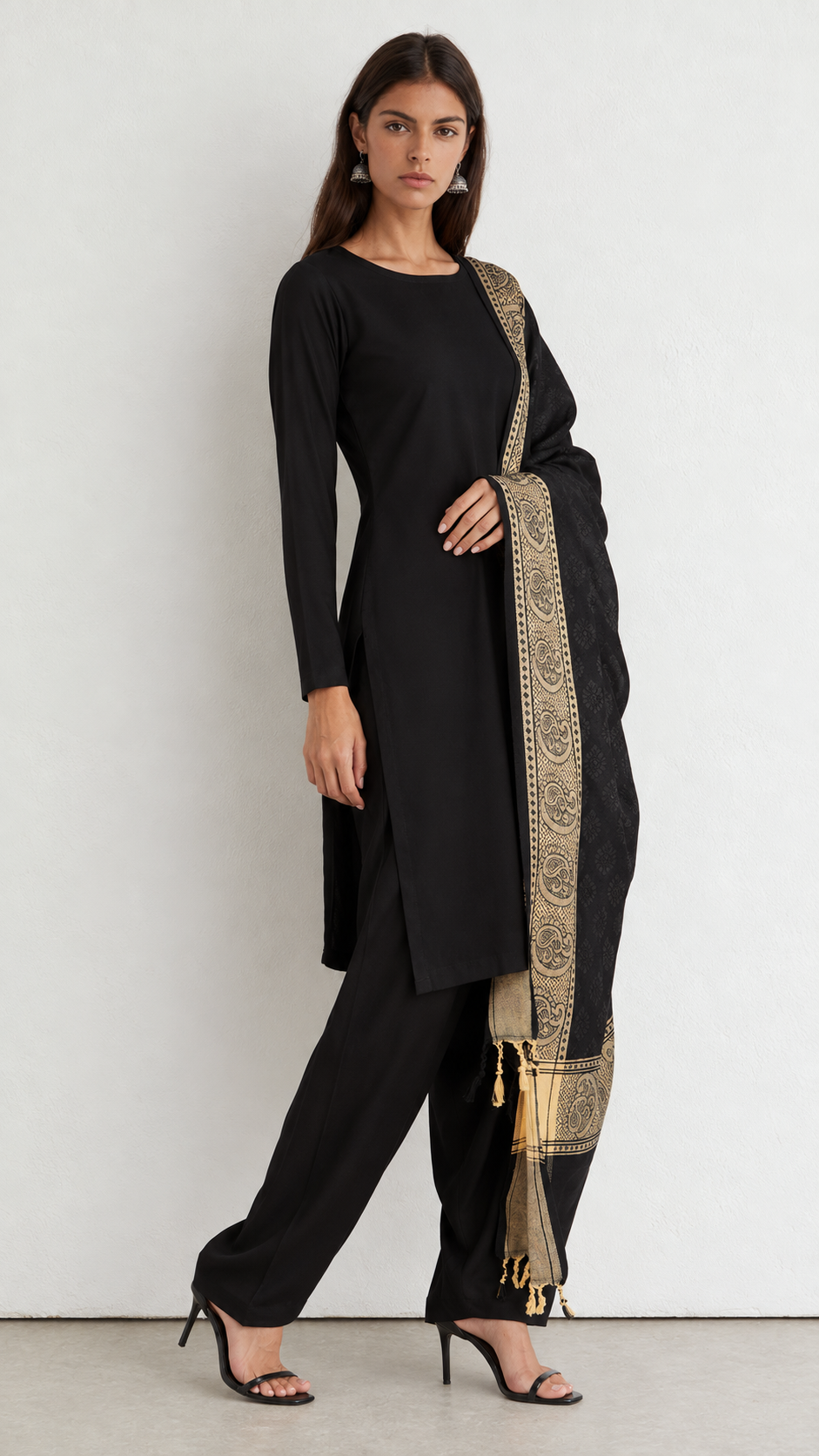 A woman modeling a black three-piece traditional outfit with a gold patterned dupatta, standing against a plain off-white wall.