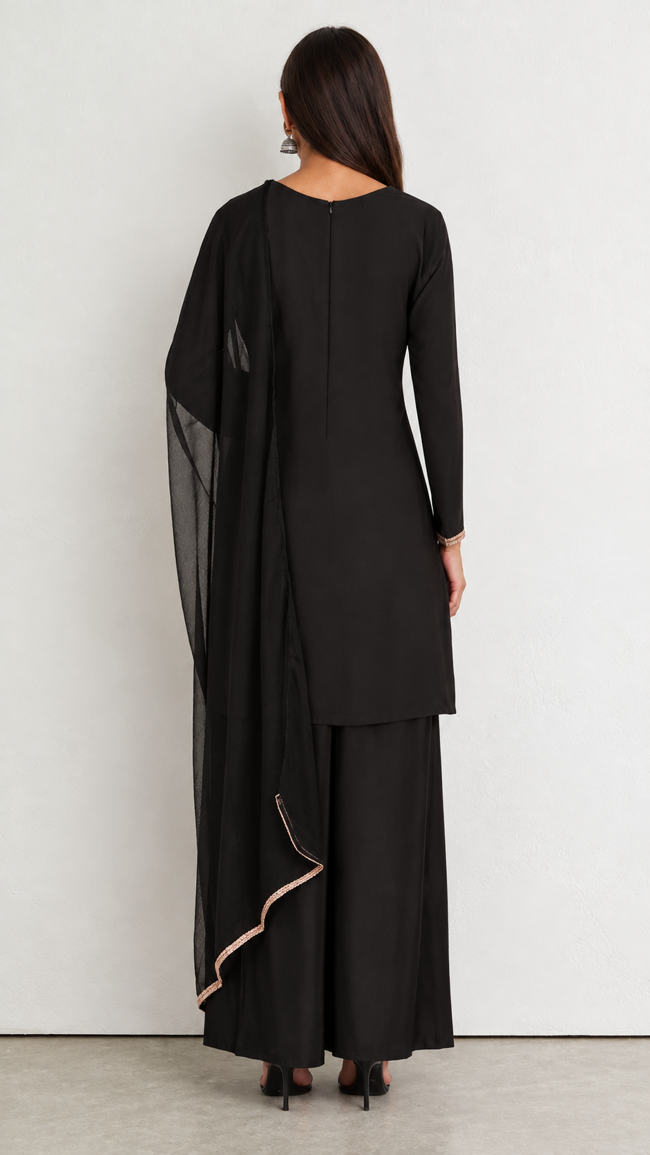 A woman shown from the back wearing a black long-sleeve kurta with matching wide-leg pants and a sheer black dupatta draped over one shoulder, against a plain light gray wall.