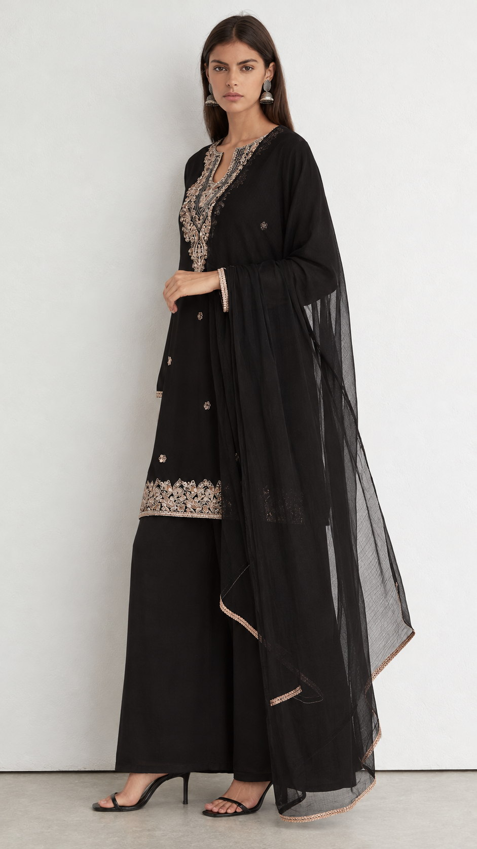 A woman models a black traditional outfit consisting of a long-sleeved embroidered kurta, wide-leg pants, and a sheer black dupatta, set against a plain light gray studio background.