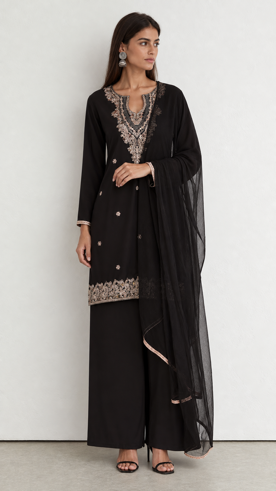 A woman modeling a black traditional outfit with intricate silver and beige embroidery on the neckline and hem, paired with wide-leg pants and a sheer black dupatta, set against a plain light gray wall.