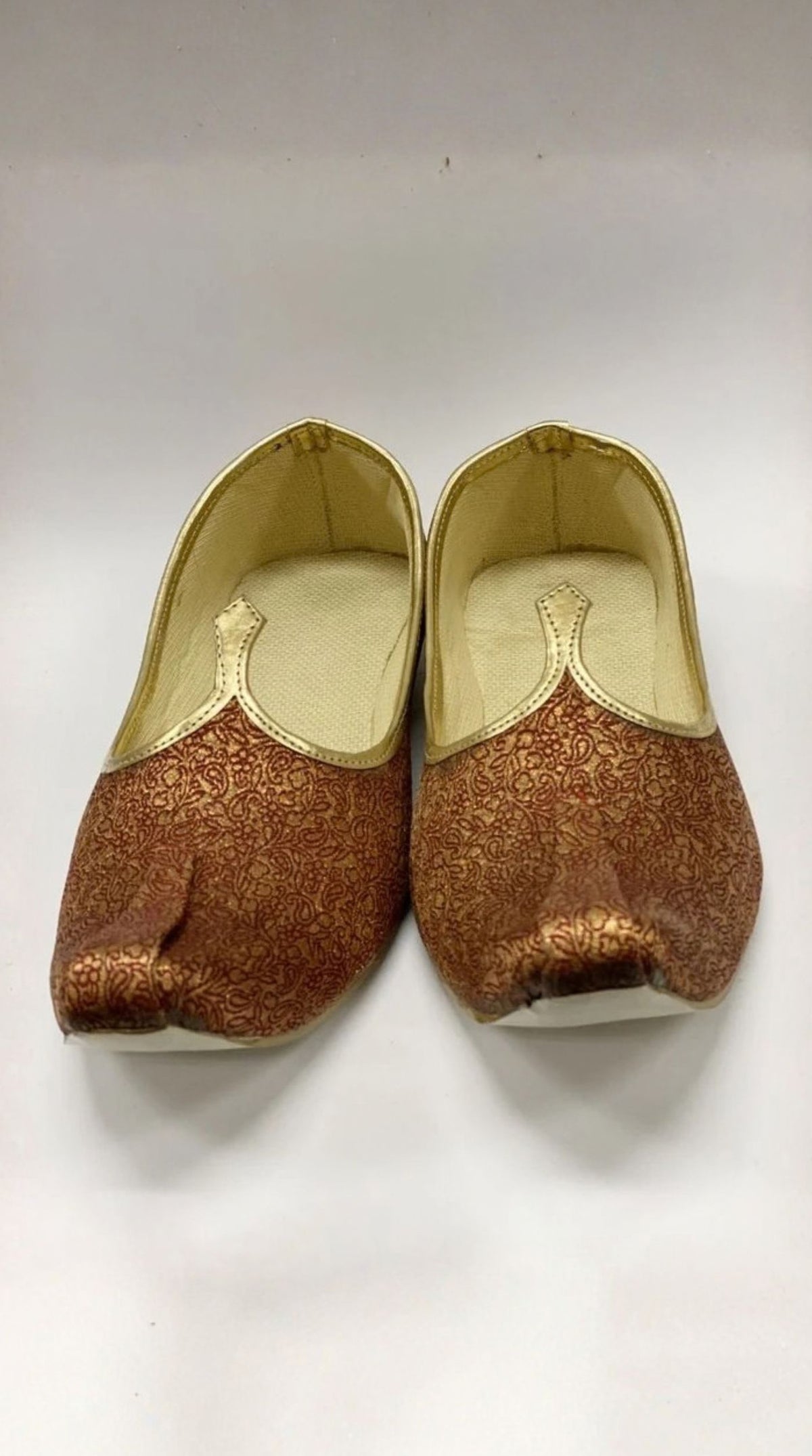 A pair of traditional Indian slip-on shoes, likely Jhumkas, featuring a metallic bronze and gold glittery fabric upper with a gold inner lining and white soles, displayed on a plain white background.