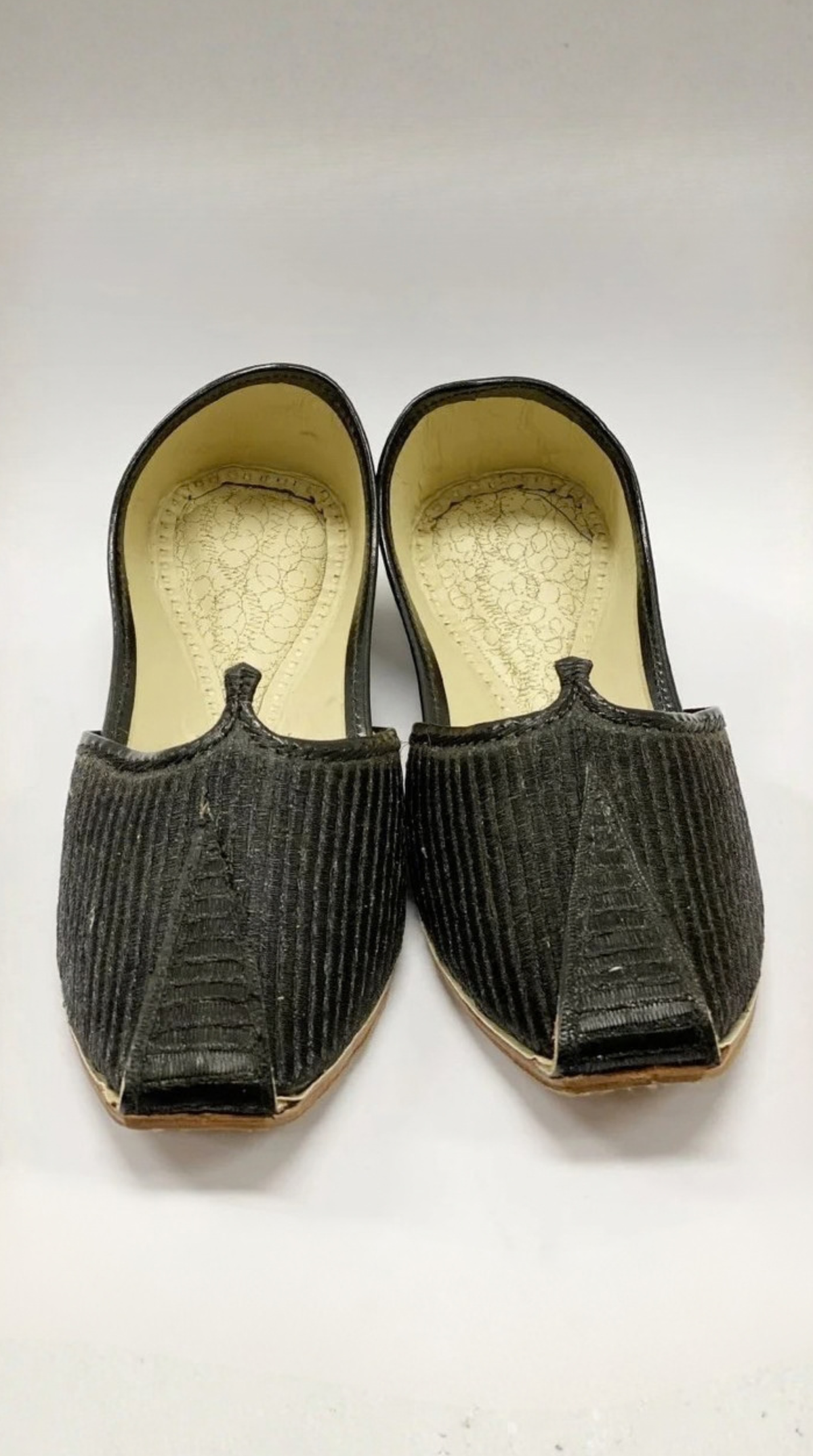 A pair of traditional black jhumka-style slip-on shoes with a textured, vertically ribbed fabric upper and a light beige insole, shown against a plain white background.