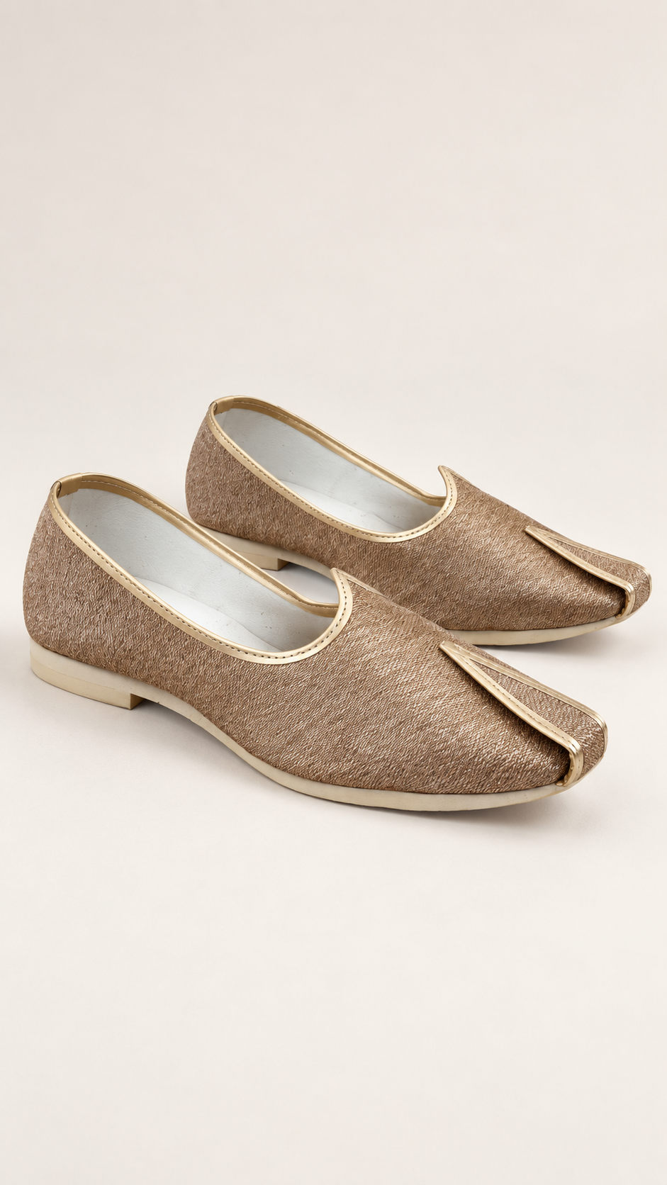 A pair of traditional Indian slip-on shoes, known as sherwani kehat, in a textured metallic gold fabric with gold trim and a pointed toe, shown on a light beige background.