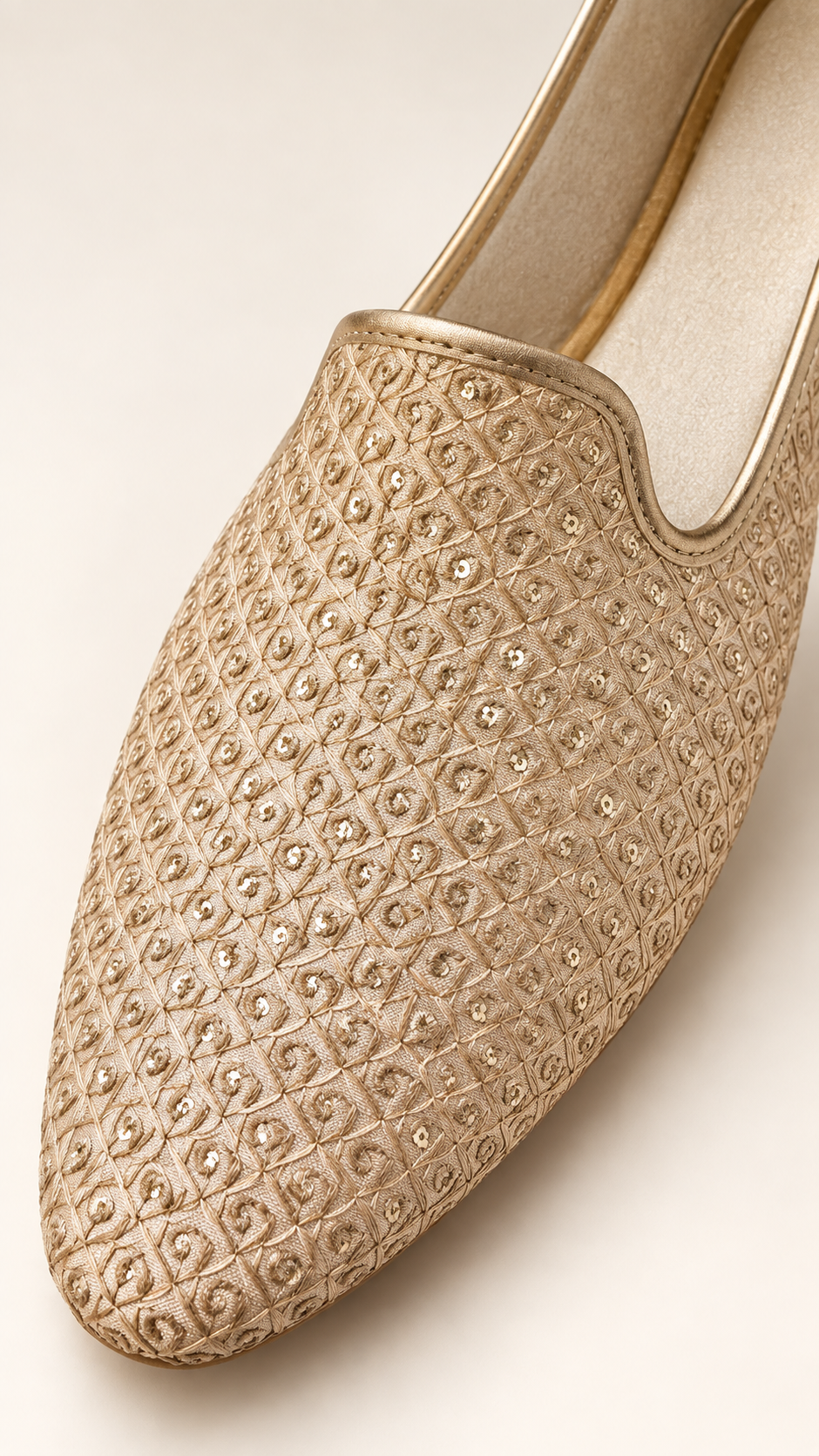 A close-up of a gold slip-on loafer featuring an intricate, textured geometric pattern with small sippy embellishments, set against a solid light beige background.
