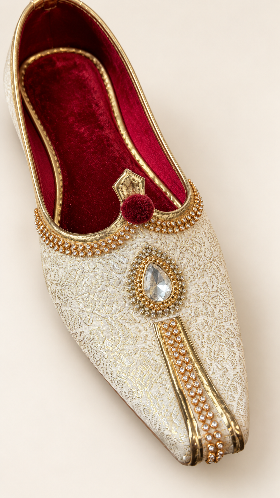 An ornate off-white embroidered Mughal-style shoe with gold trim, a red velvet interior, and a decorative jeweled toe ornament, shown on a light beige background.