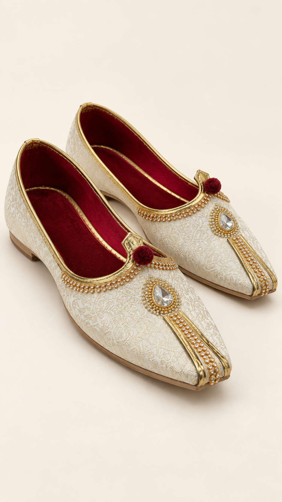 A pair of traditional Indian groom's shoes (Mithai Khaddar) in off-white brocade fabric with gold embroidery, red velvet lining, and a clear rhinestone embellishment on the toe.
