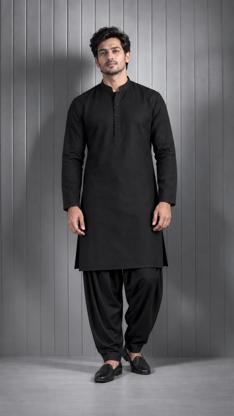 A man modeling a black long-sleeve kurta with a buttoned collar, paired with black pants and black loafers, standing against a gray paneled studio wall.