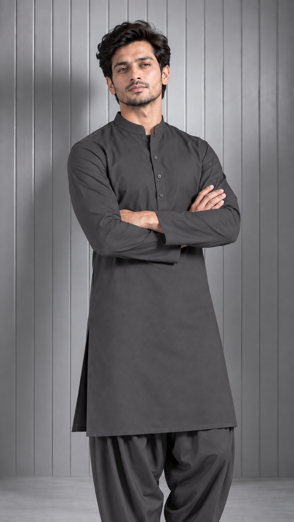 A man wearing a dark gray long-sleeve kurta and matching pants, standing with arms crossed against a gray paneled studio background.