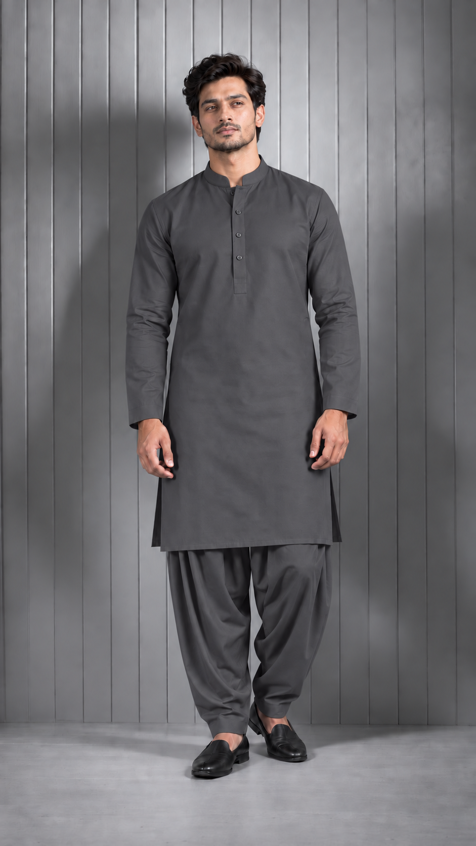 A man modeling a dark gray long-sleeve kurta with a mandarin collar and button placket, paired with matching dark gray salwar pants and black loafers, standing against a gray paneled studio backdrop.