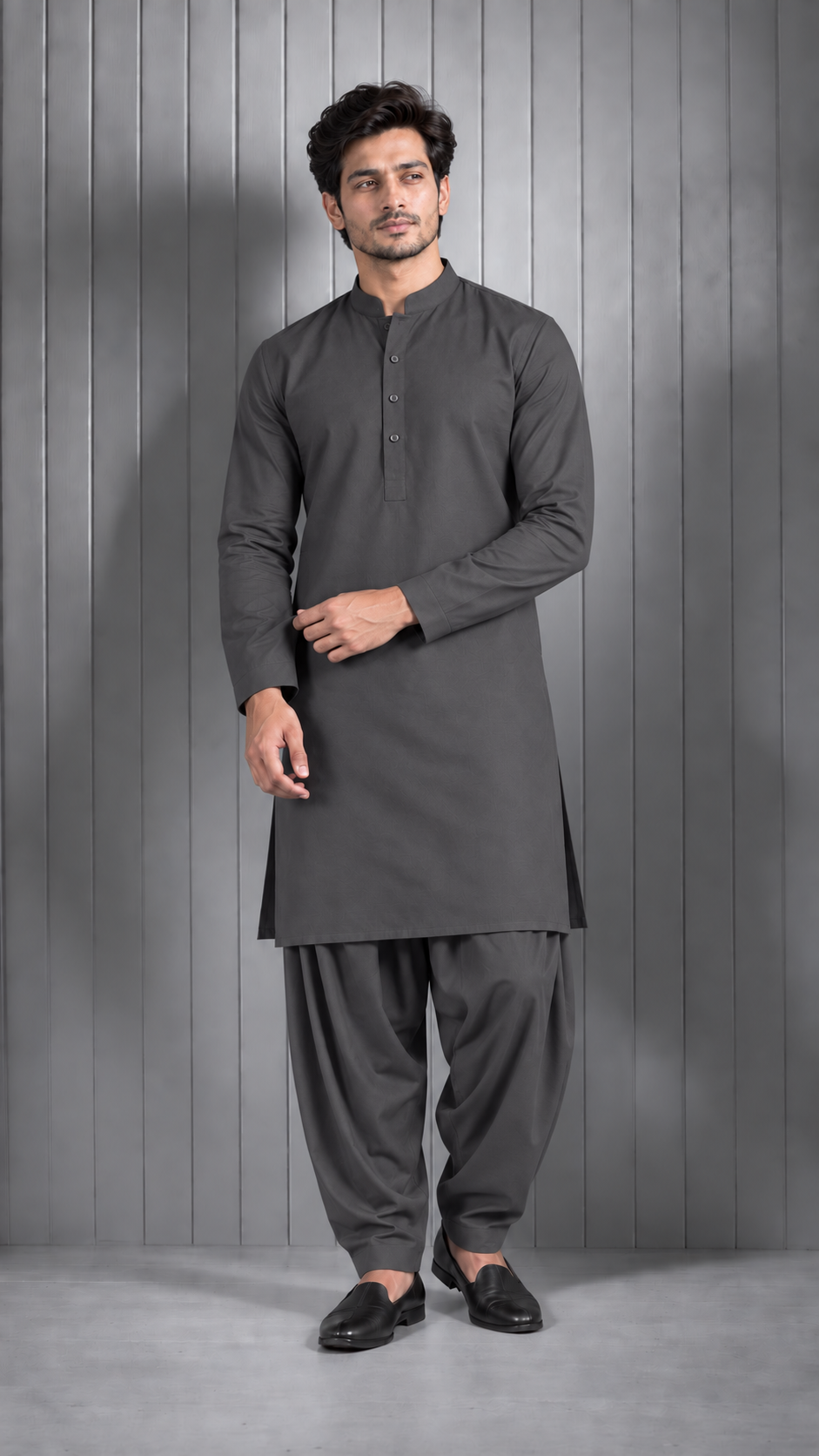 A man modeling a dark gray long-sleeve kurta with a mandarin collar and front buttons, paired with matching dark gray salwar pants and black loafers, standing against a gray paneled studio backdrop.