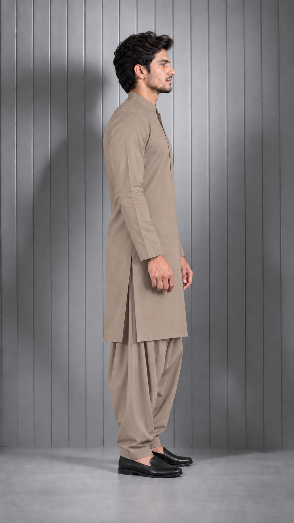 A man wearing a light brown traditional shirtdress (kurta and shalwar set) standing in profile against a gray paneled studio wall.