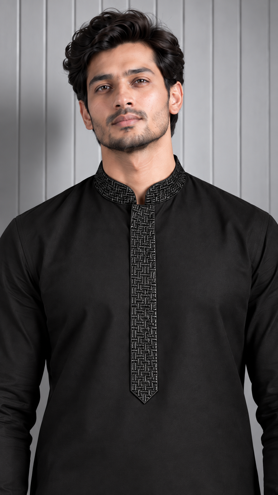 A man wearing a black long-sleeve kurta with a textured geometric pattern on the collar and placket, standing against a gray paneled background.