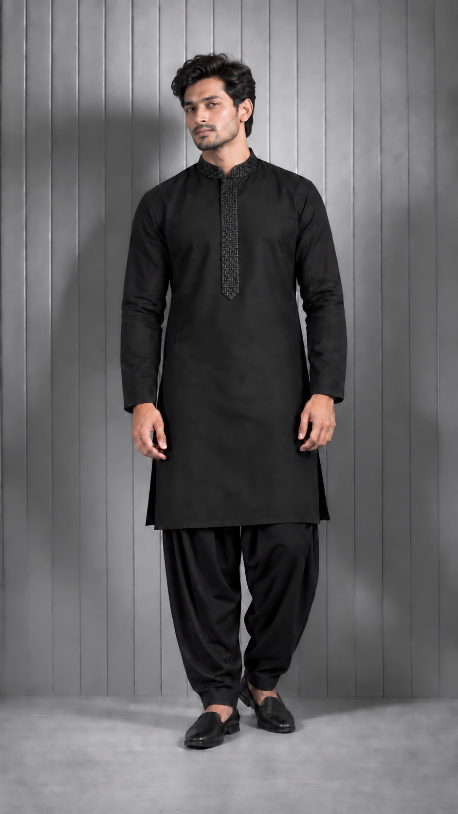 A man modeling a black long-sleeve kurta with a patterned black collar trim, paired with black pants and black slip-on shoes, standing against a gray paneled studio background.