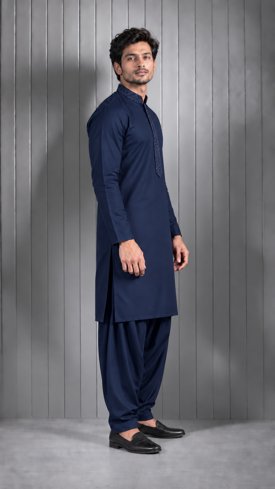 A man modeling a navy blue traditional Indian outfit consisting of a long-sleeve kurta and matching pants, standing against a gray paneled studio wall.